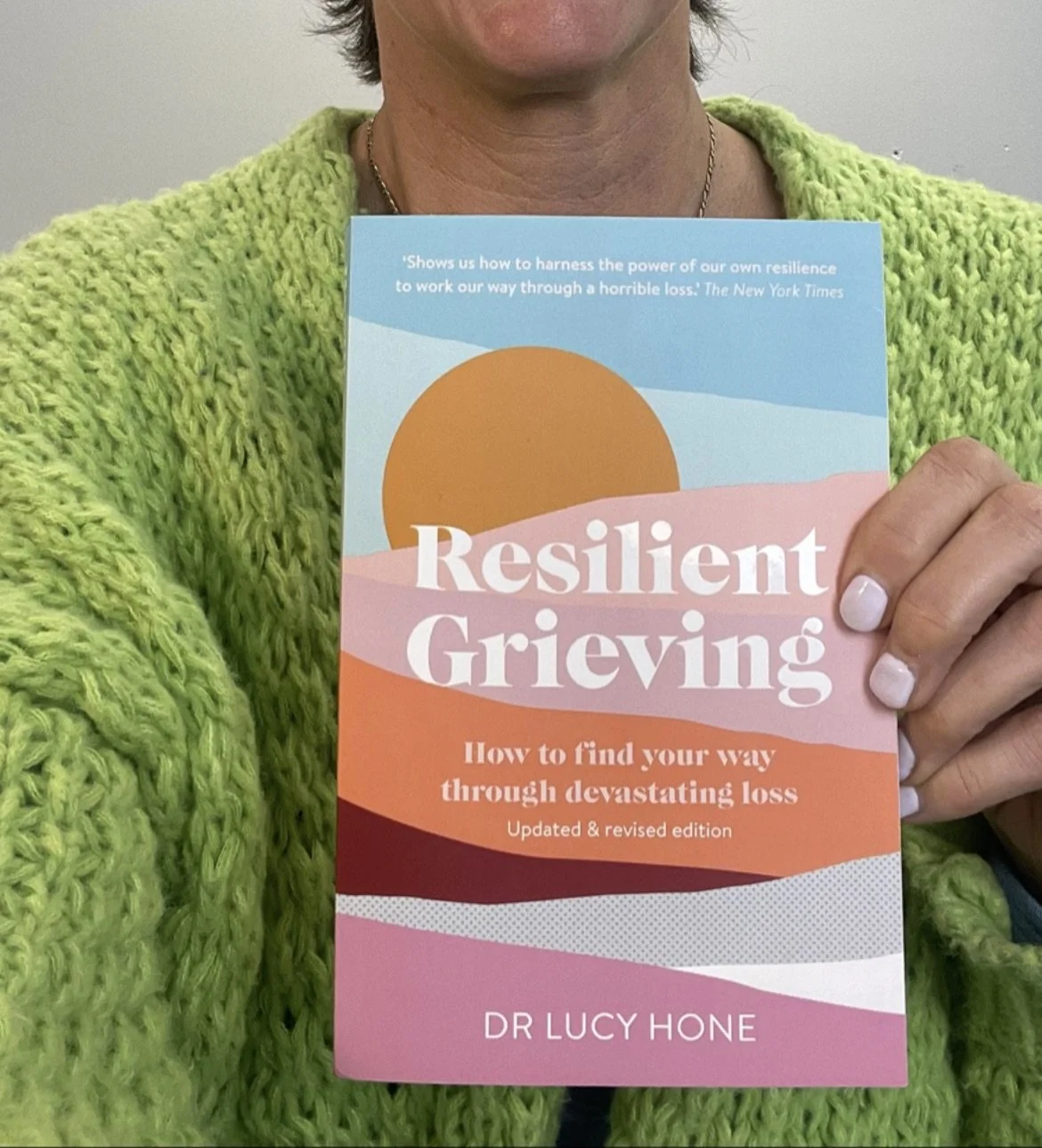 Person holding a book titled "Resilient Grieving" by Dr. Lucy Hone, wearing a bright green knitted sweater.