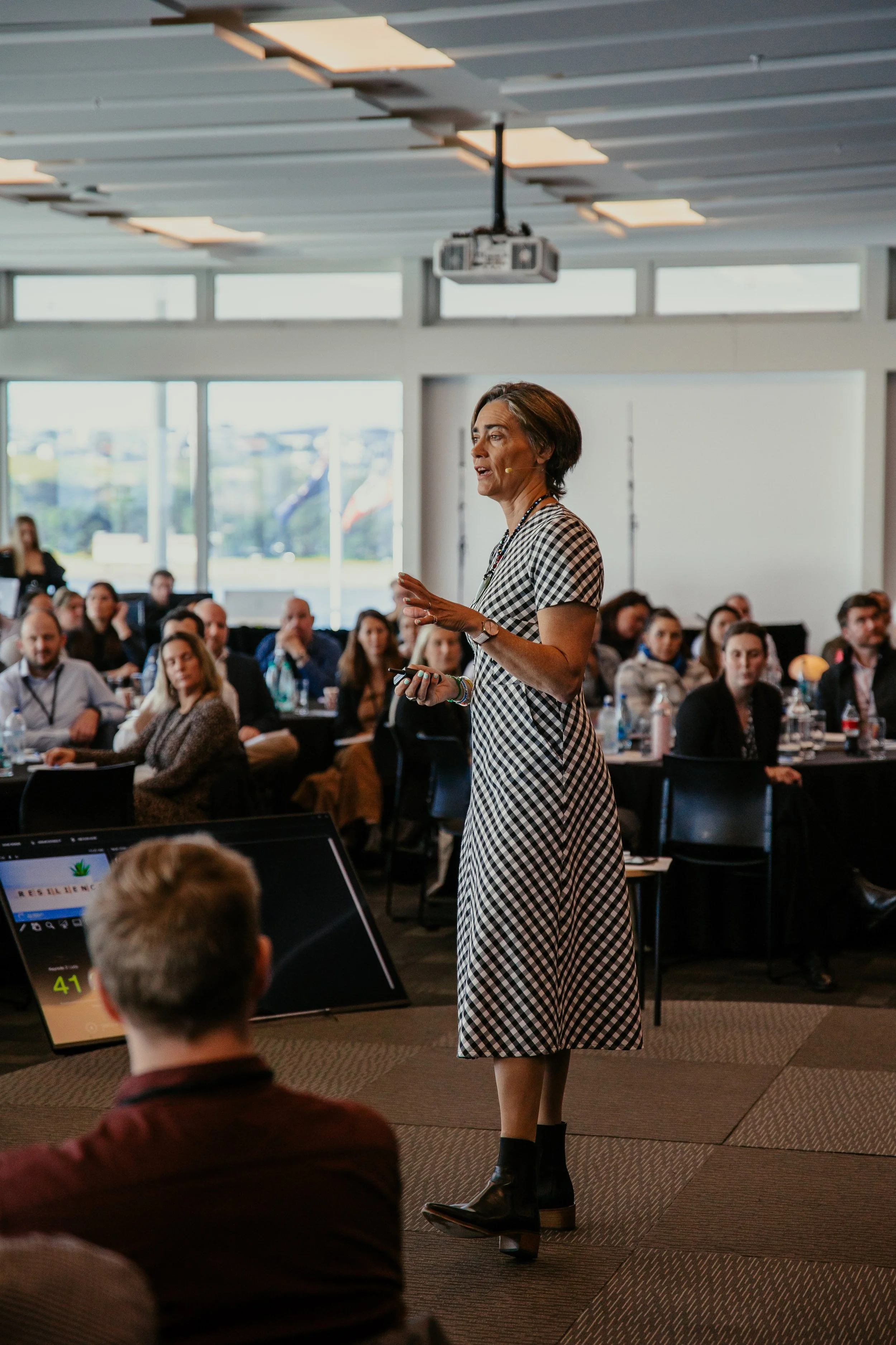 Dr Lucy Hone, in a checkered dress, giving a presentation to a seated audience in a conference room at a corporate event.