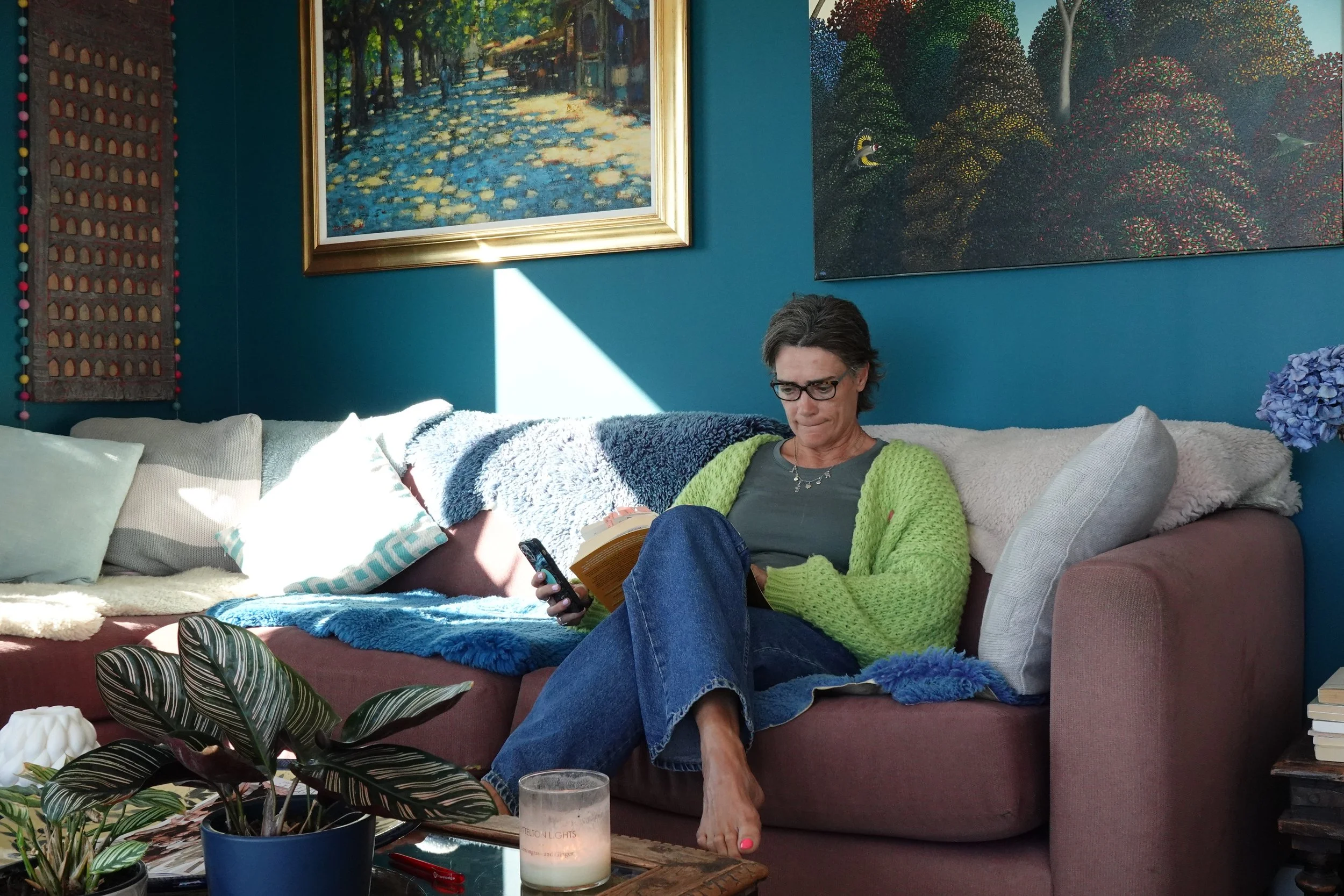 Lucy relaxing on a pink sofa, reading in a colourful living room surrounded by art, books, and soft furnishings.