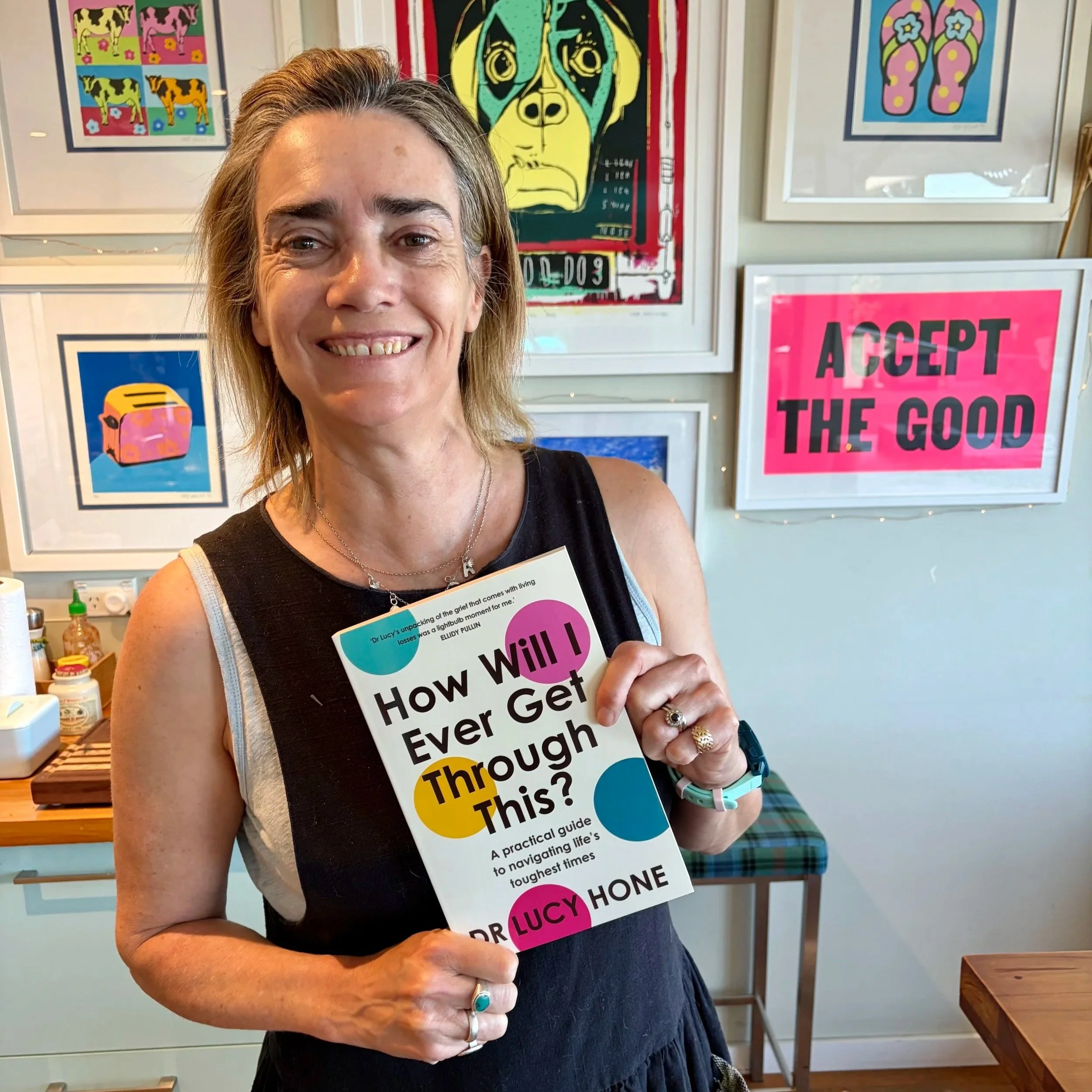 A woman with shoulder-length blonde hair holding a book titled "How Will I Ever Get Through This?" in a room decorated with colorful framed artwork, including a sign that says "ACCEPT THE GOOD."