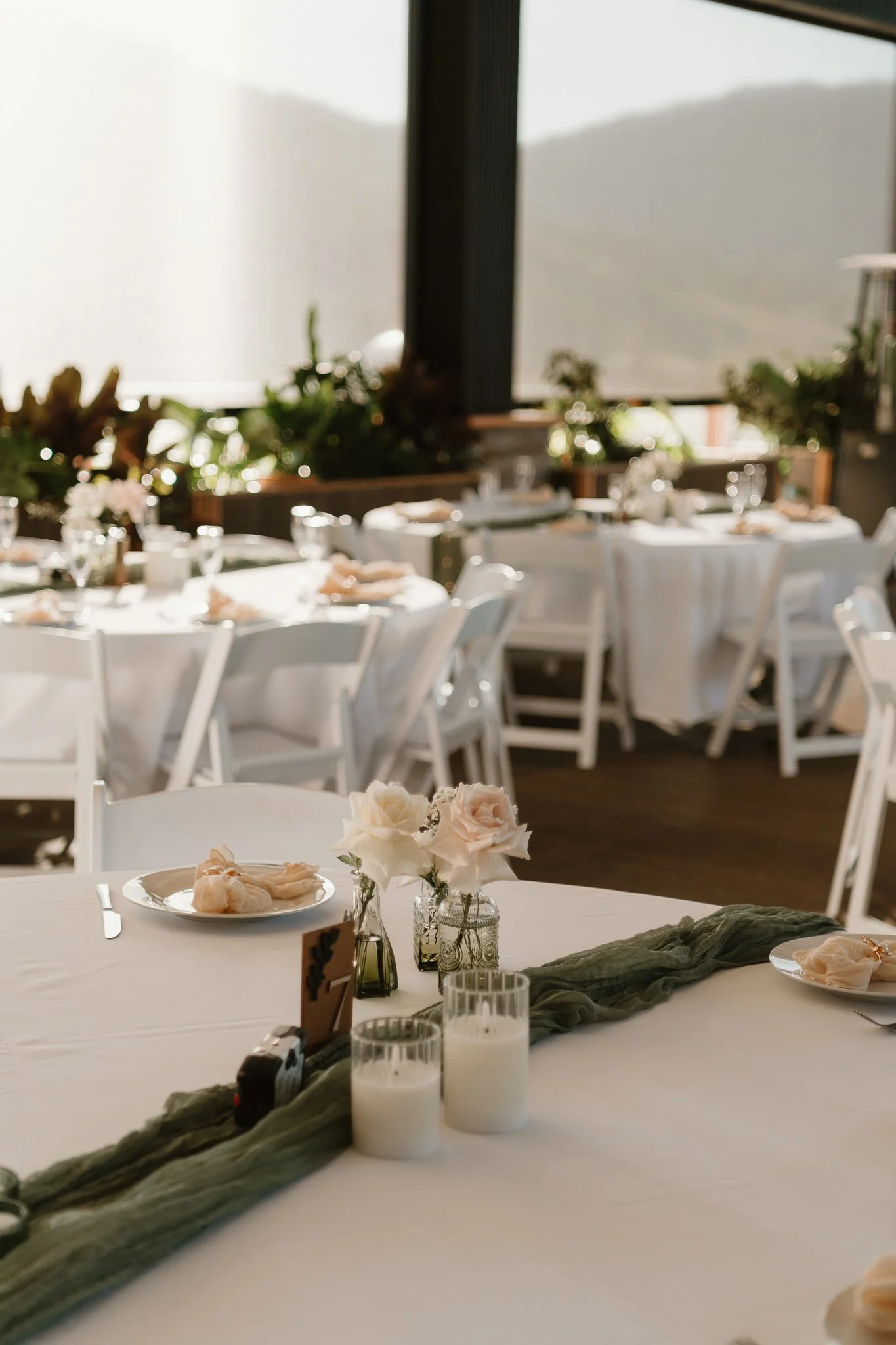 Wedding reception setup with tables decorated with flowers, candles, and elegant tableware, set in a room with large windows and mountain views.