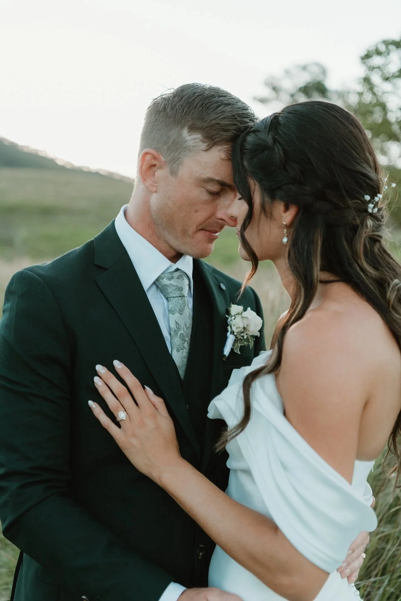 A couple dressed in wedding attire embracing outdoors during daytime with their foreheads touching, eyes closed, and hands on each other's waist.