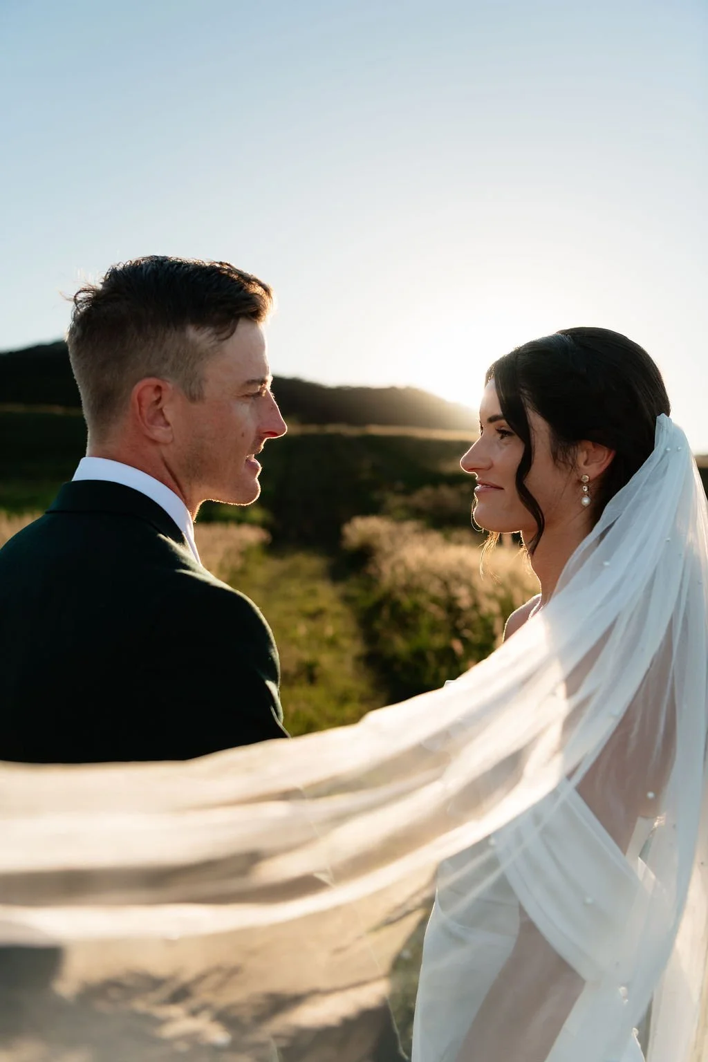 A bride and groom facing each other outdoors during sunset, with the bride wearing a white wedding gown and veil, and the groom in a black tuxedo, against a natural landscape background.