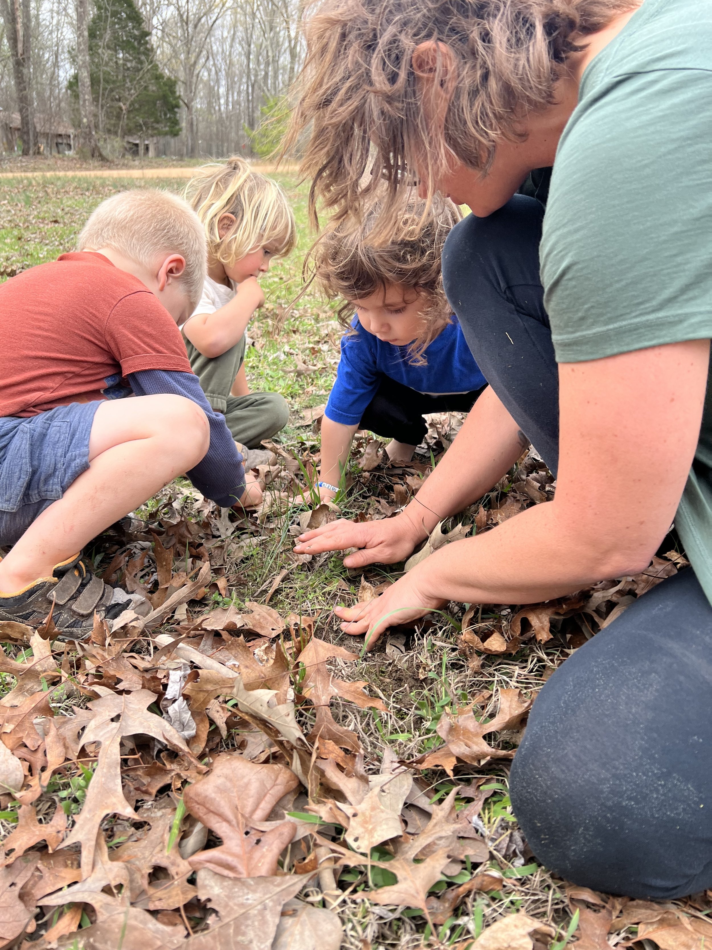 A group of children and an adult crouching on the ground in a forested area, exploring leaves and soil together.