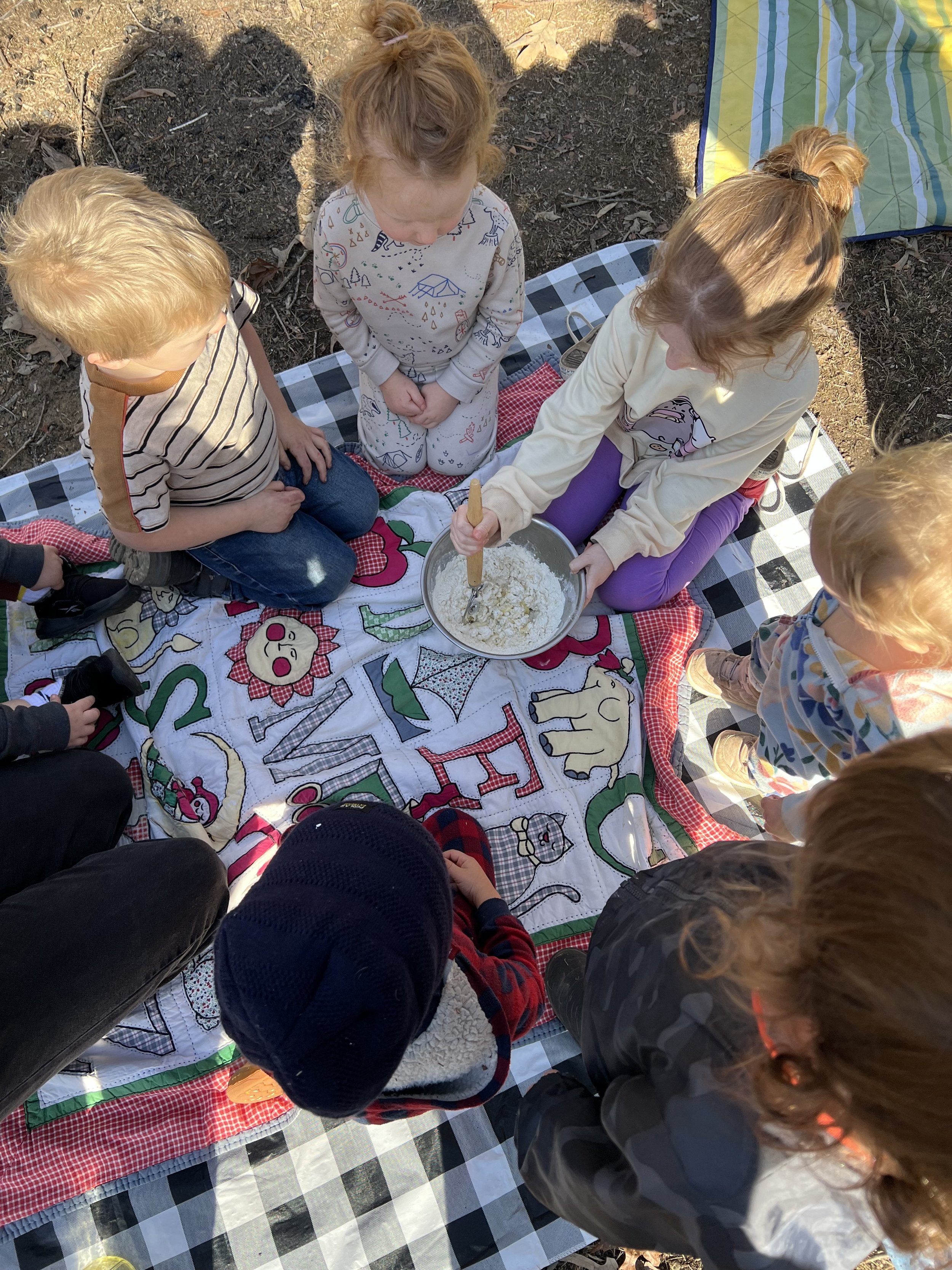 Group of children kneeling on a picnic blanket with one child stirring a bowl of ingredients outdoors.