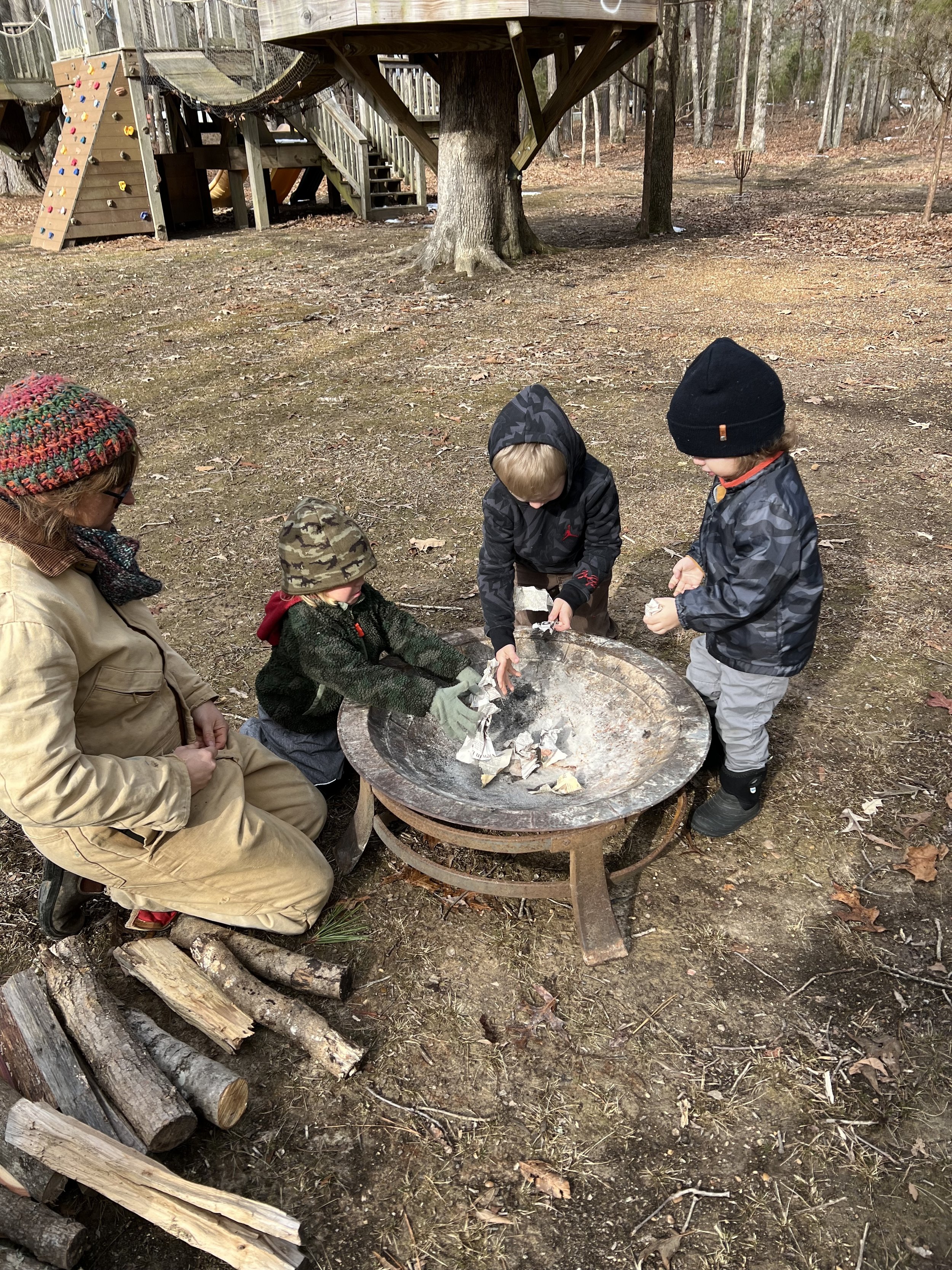 Three children and an adult engaging in outdoor activity near a fire pit, surrounded by trees and a pile of wood, with a wooden playground in the background.