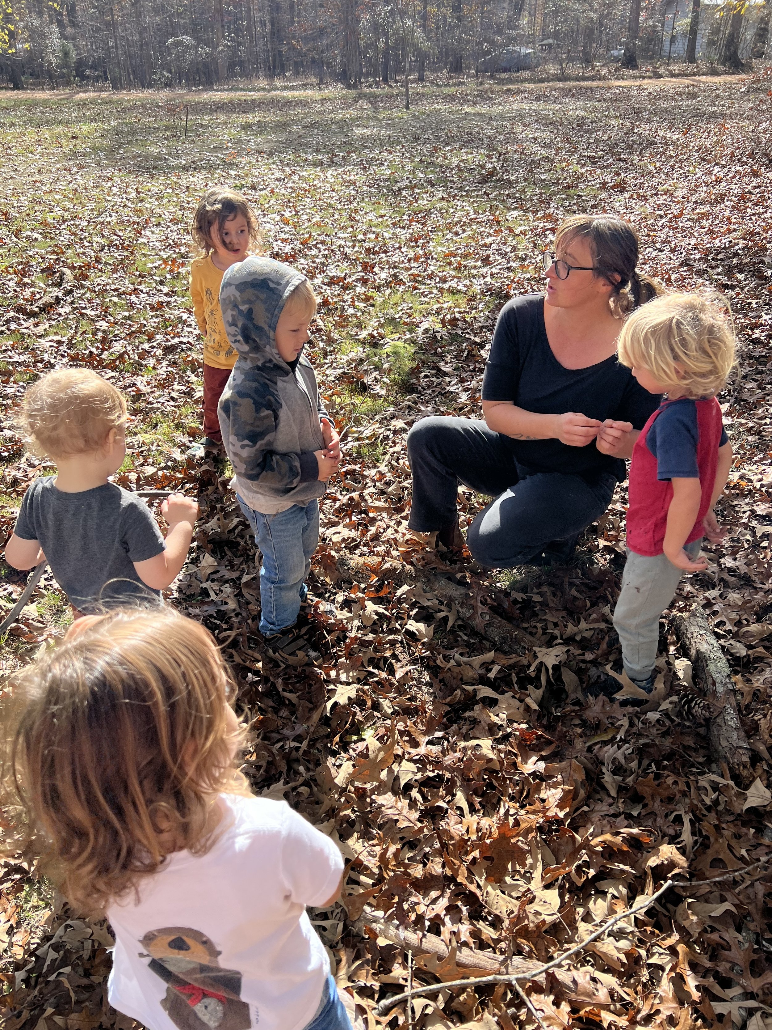A group of children and an adult are gathered in a forest clearing surrounded by fallen autumn leaves. The adult, wearing glasses and a black top, is kneeling and interacting with the children, who wear casual clothes and seem engaged. The background