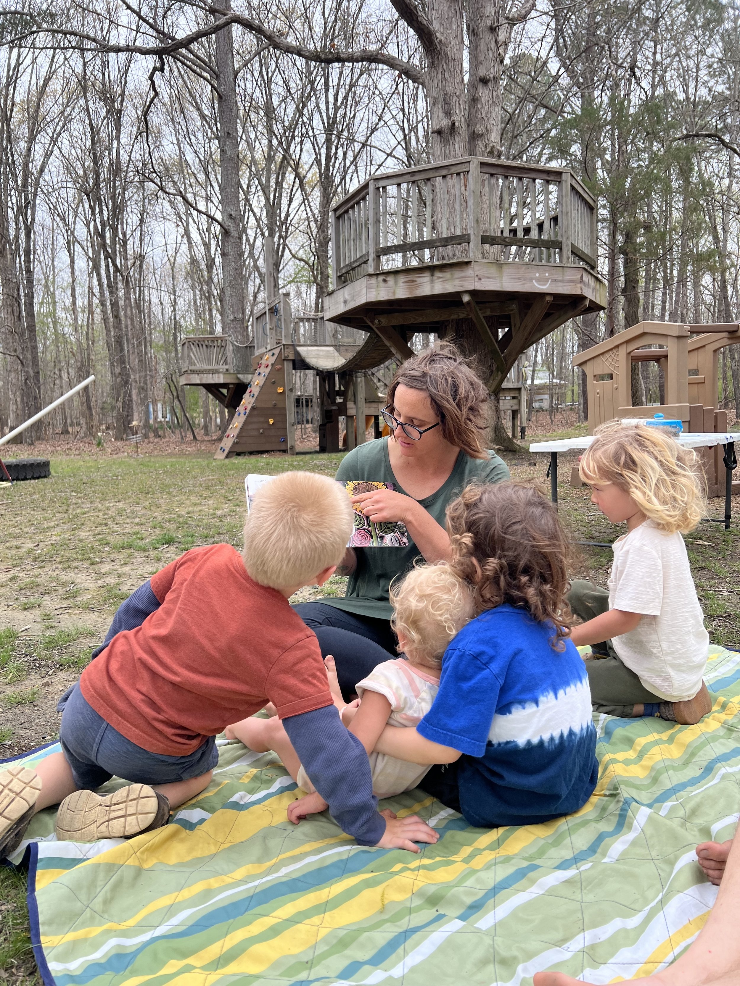 A woman sitting outdoors on a picnic blanket with a group of children, reading a book to them. In the background, a wooden treehouse and playground are visible among tall trees.