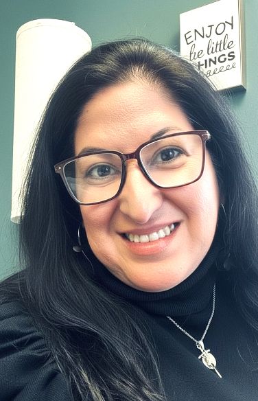 A woman, with glasses, smiling and standing against a green wall.