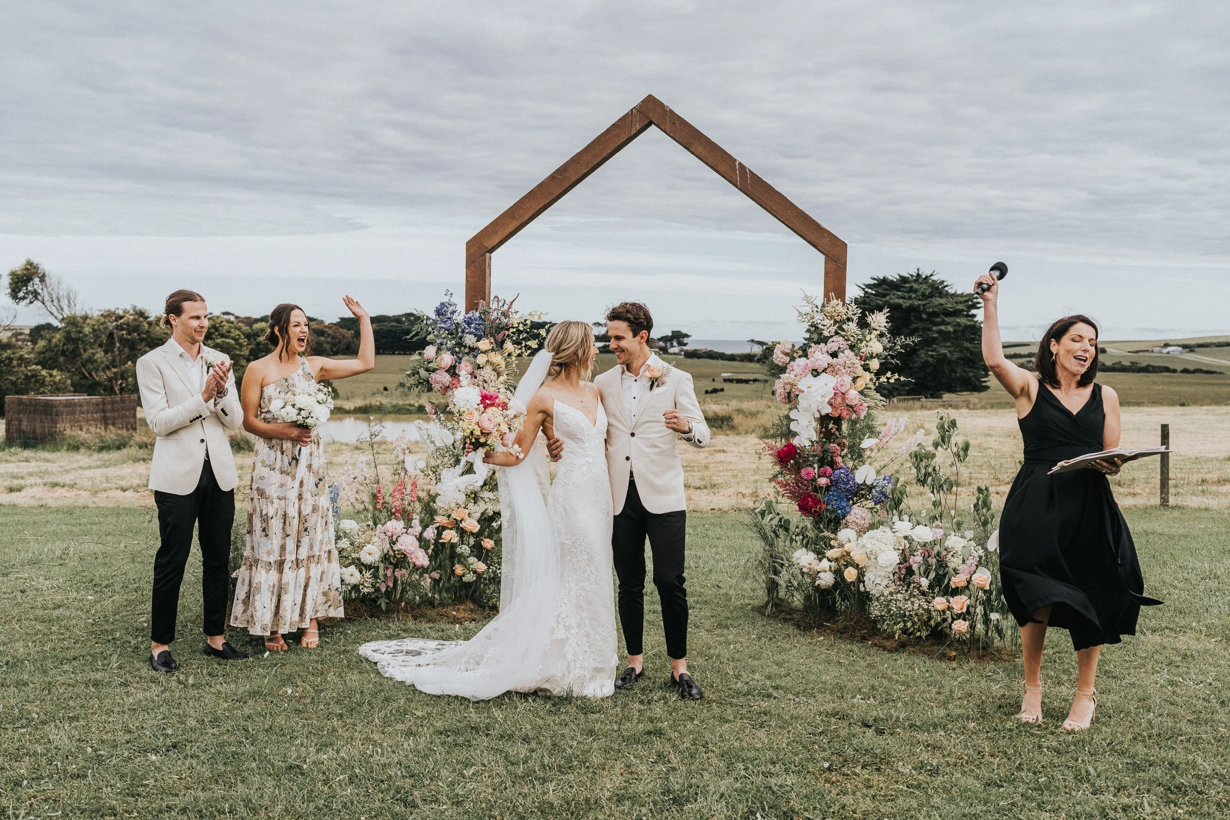 Outdoor wedding ceremony with a couple kissing, celebrant raising hand, guests celebrating, floral arch and scenic background.