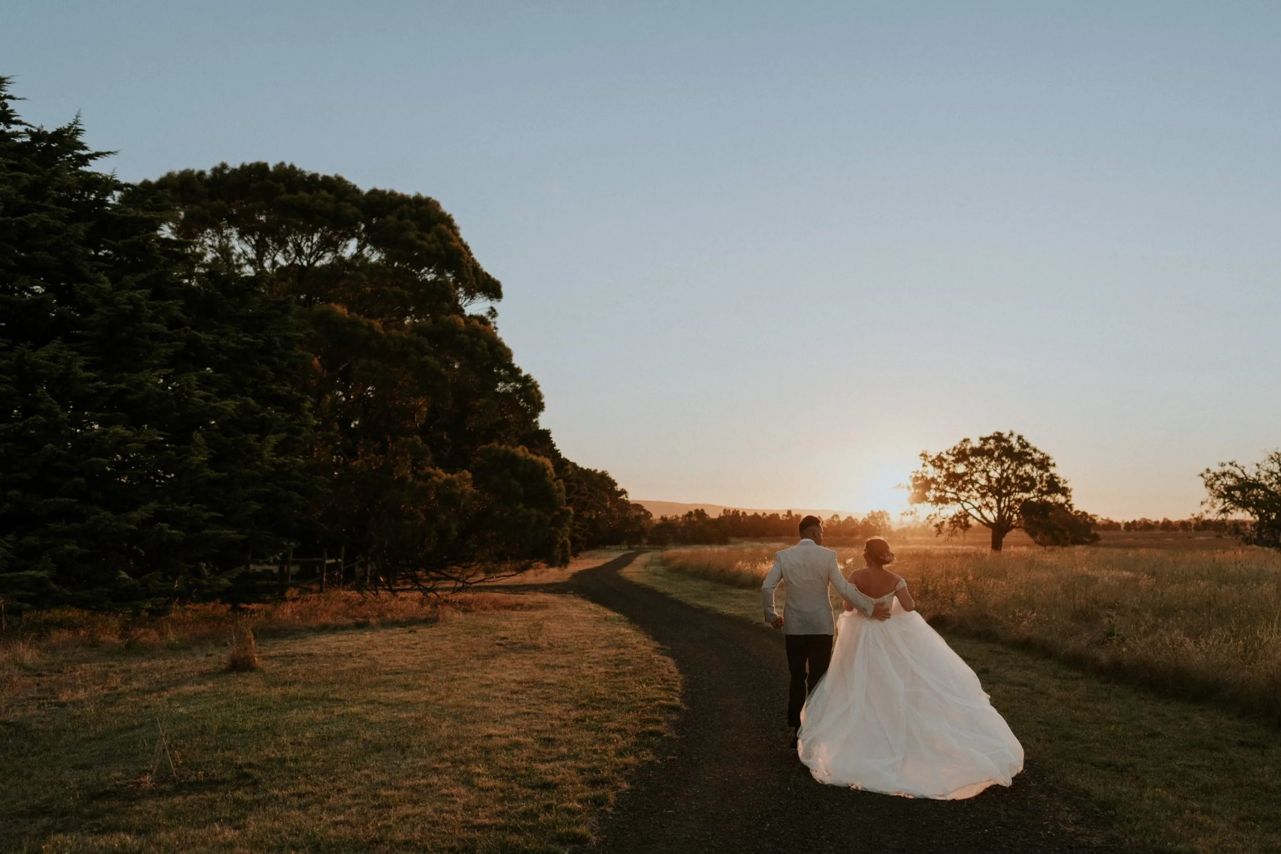 A couple in wedding attire walking hand in hand on a dirt path through a countryside landscape at sunset, with trees on either side and a clear sky.