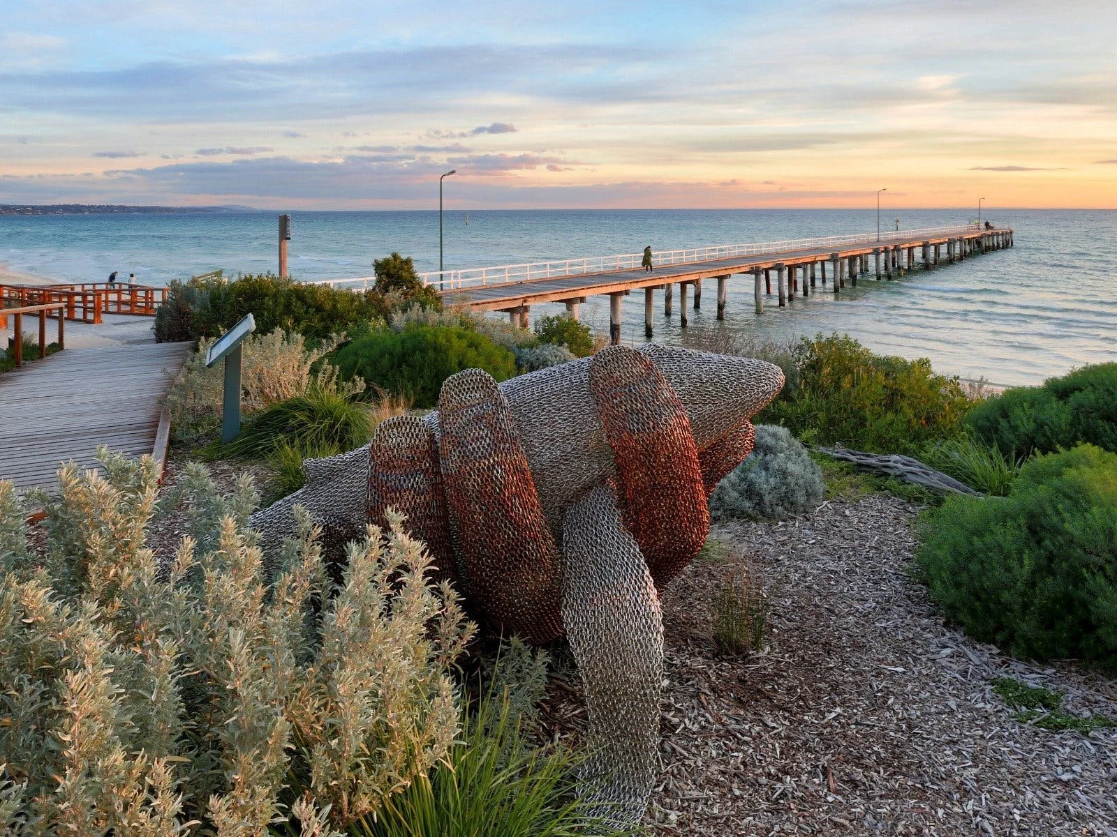 A coastal scene with a wooden pier extending into the ocean at sunset, surrounded by green bushes and a sculpture of interconnected metal rings in the foreground.