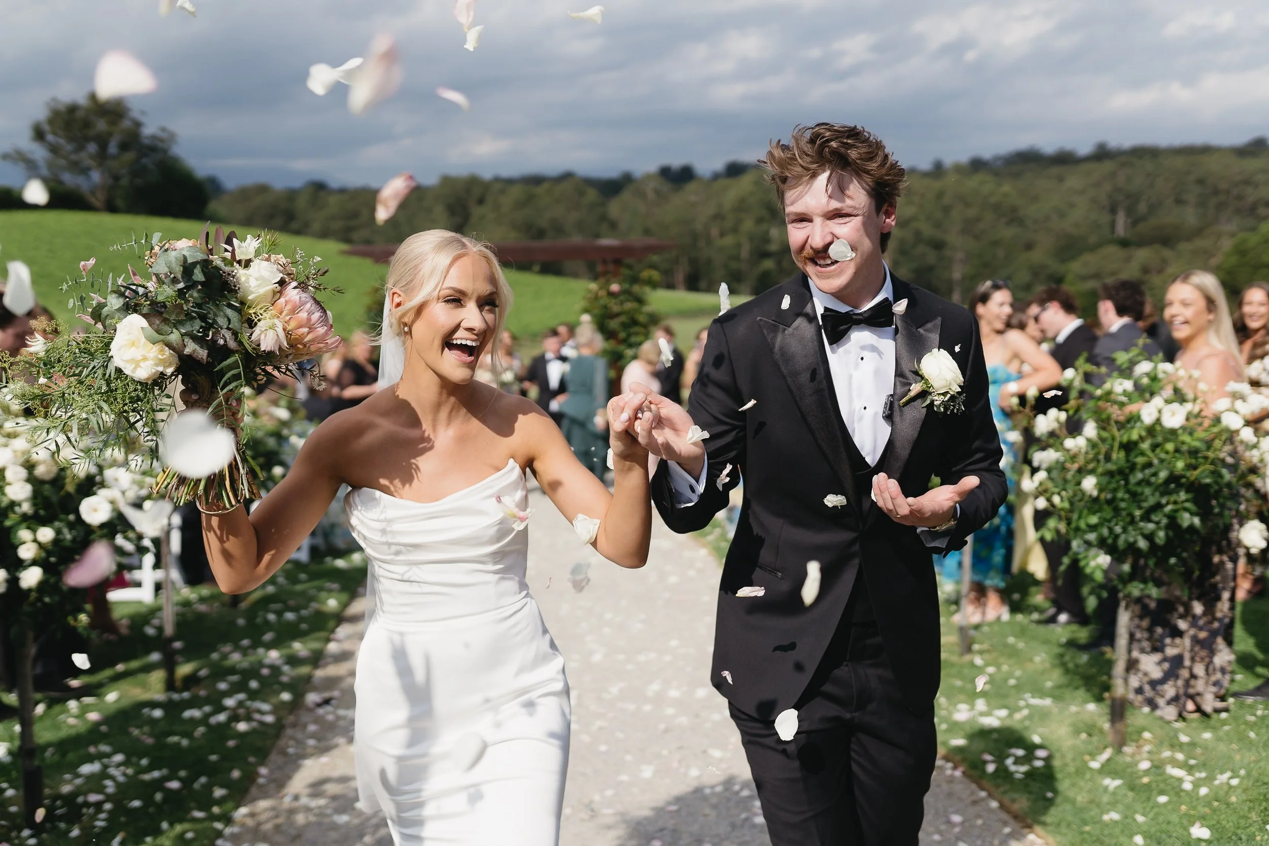 Wedding couple walking down aisle outdoors, smiling with flower petals in the air.