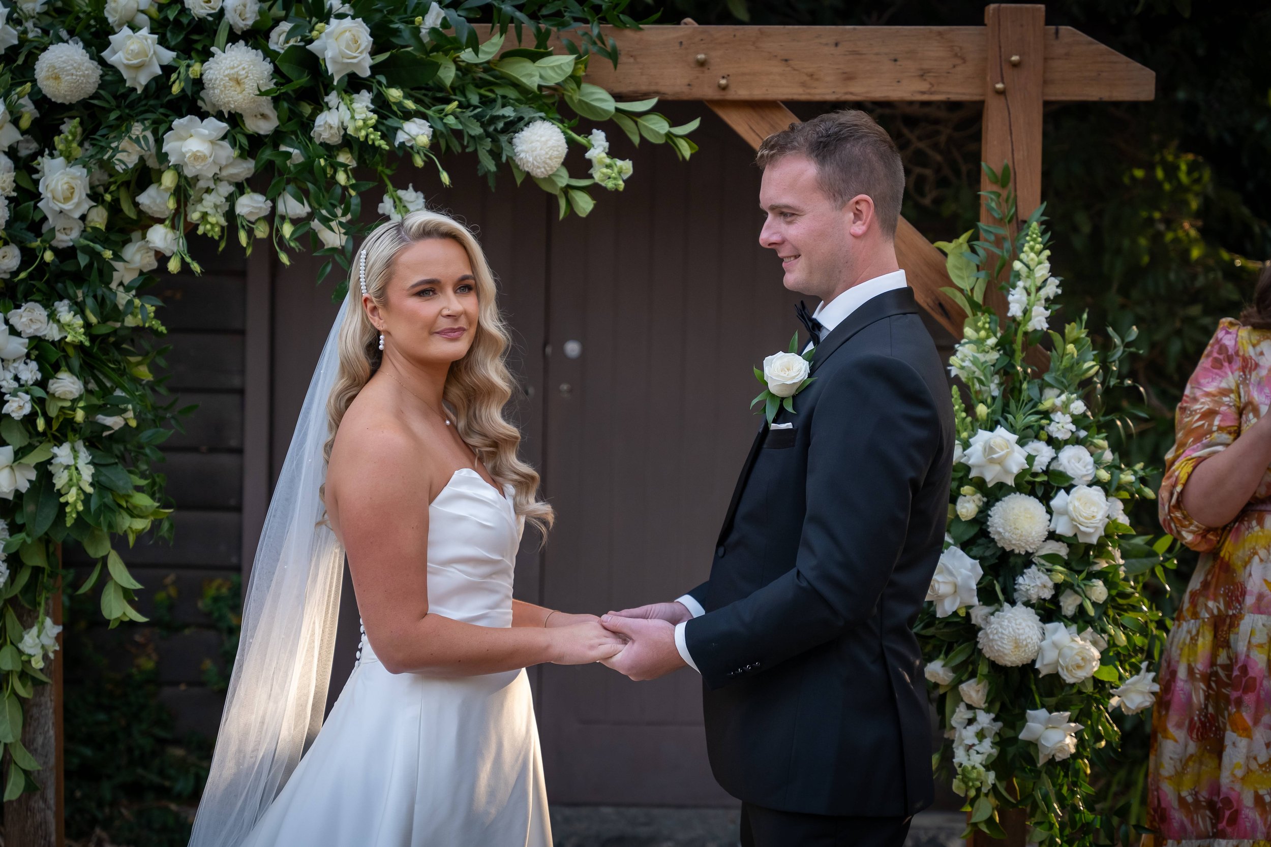 Bride and groom holding hands during a wedding ceremony under a floral arch, with white and green flower arrangements.