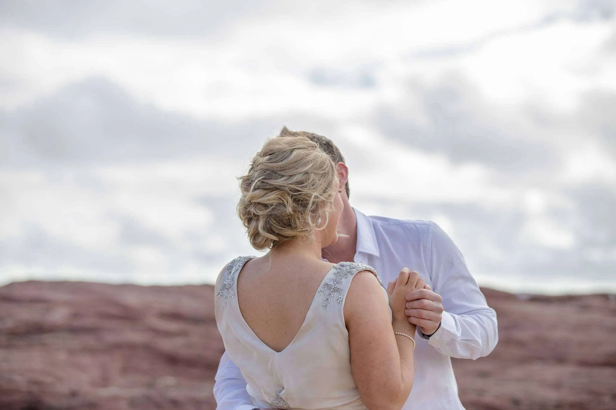 A couple kissing outdoors, woman with an updo and decorative dress, man in a white shirt, with a cloudy sky background.