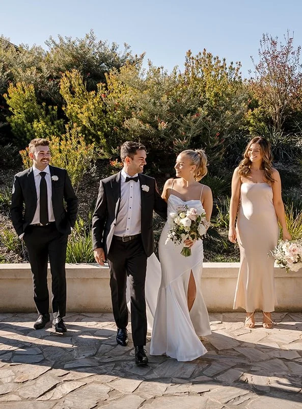 Wedding party walking outdoors with bride in white gown, groom in suit, and bridal party members holding bouquets.