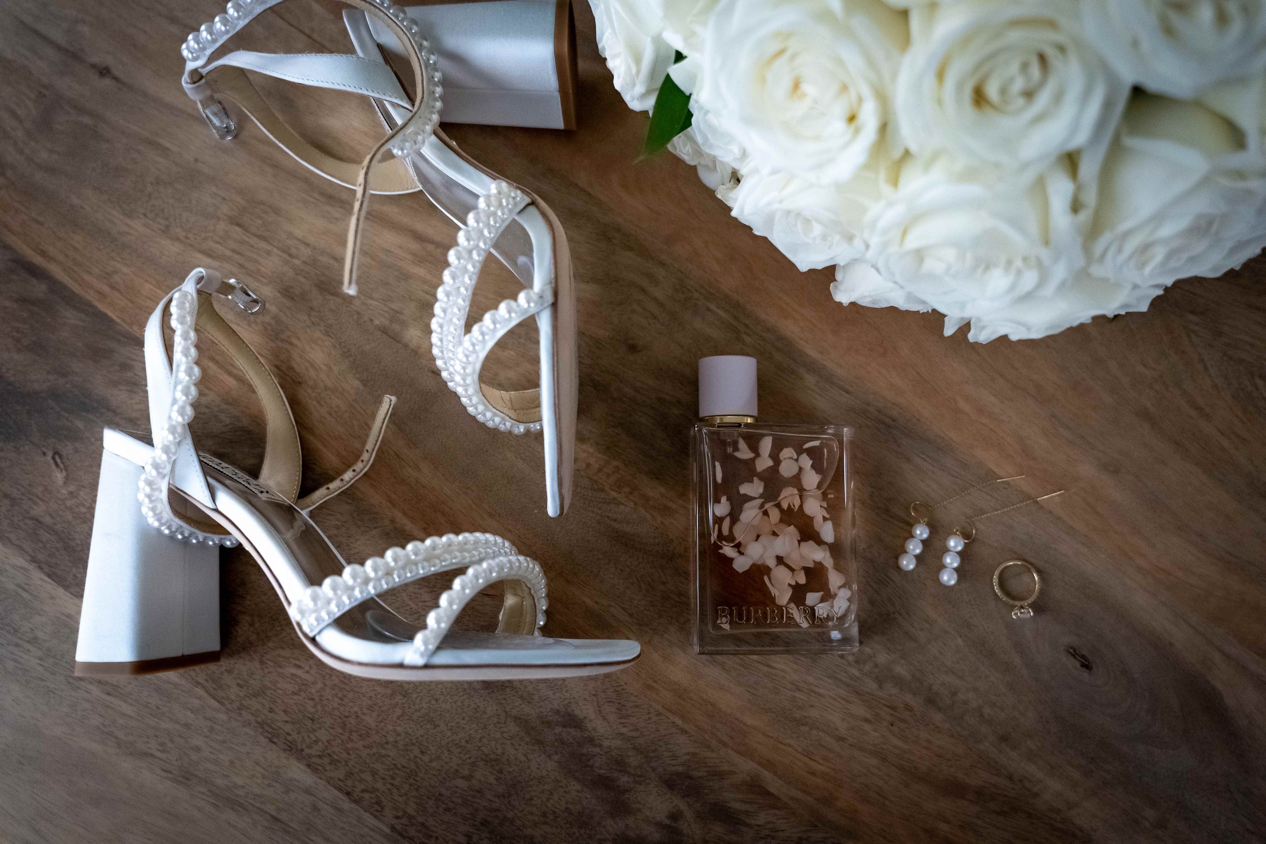 White high-heeled sandals with pearl embellishments, a bouquet of white roses, a bottle of Burberry perfume, pearl earrings, and a ring with a gemstone on a wooden surface.