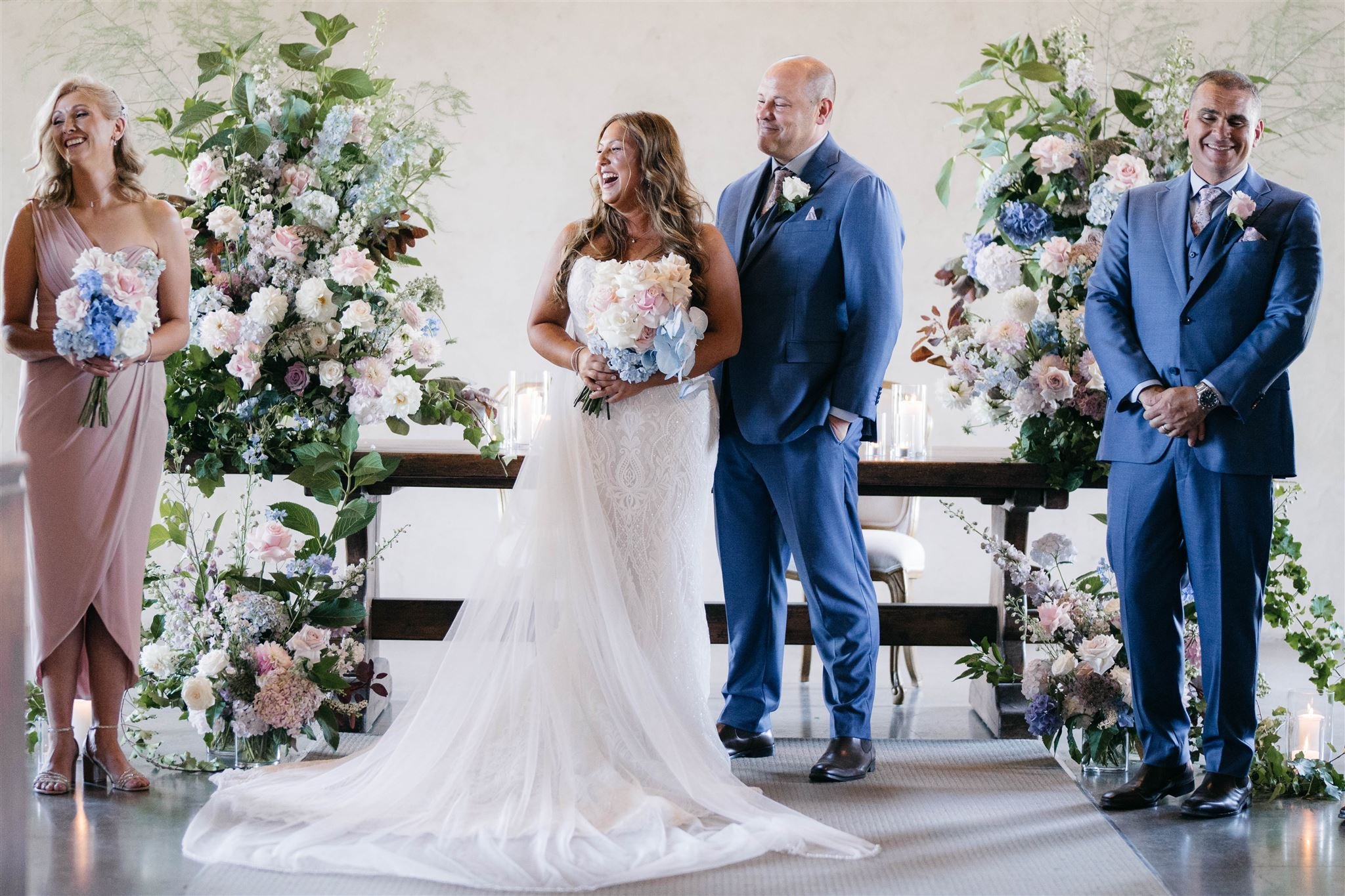 Bride and groom with wedding party in formal attire, standing before floral arrangements.