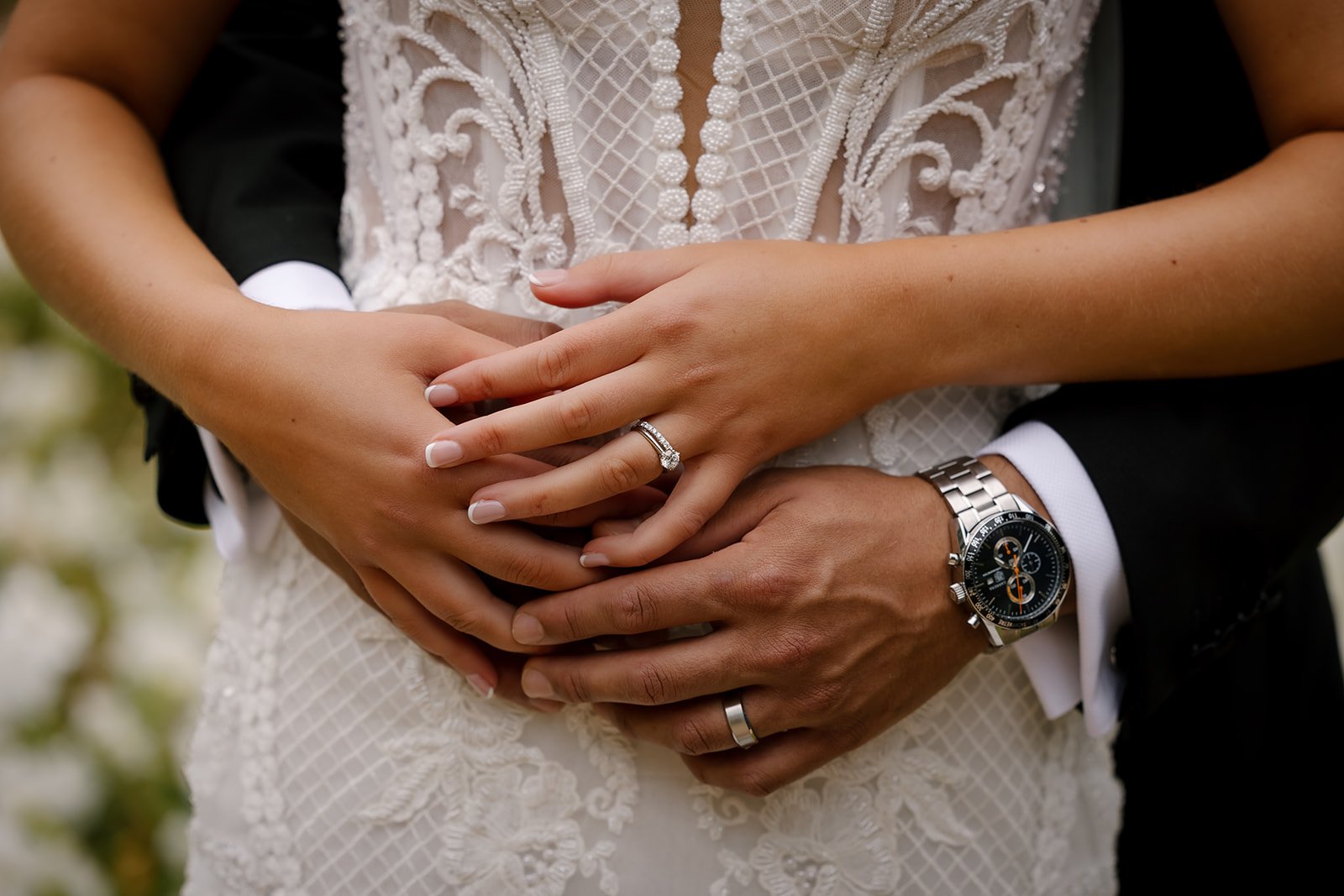 Close-up of a couple's hands showing wedding rings and a watch, with intricate lace dress detail.