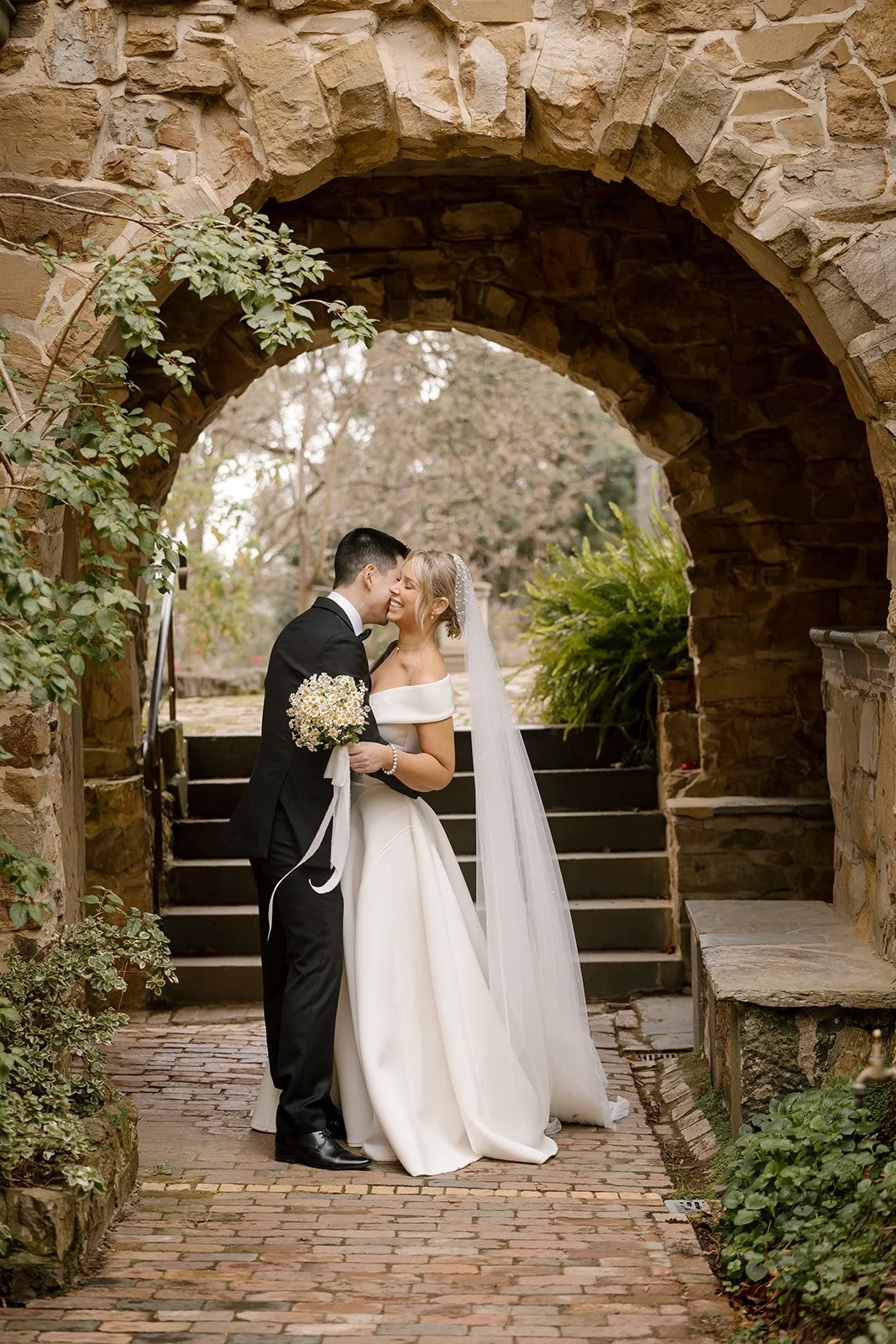 Bride and groom walking on a sandy beach at sunset, with the groom wearing a white tuxedo jacket and black pants, and the bride in a white dress with a slit. They are smiling and looking at each other.