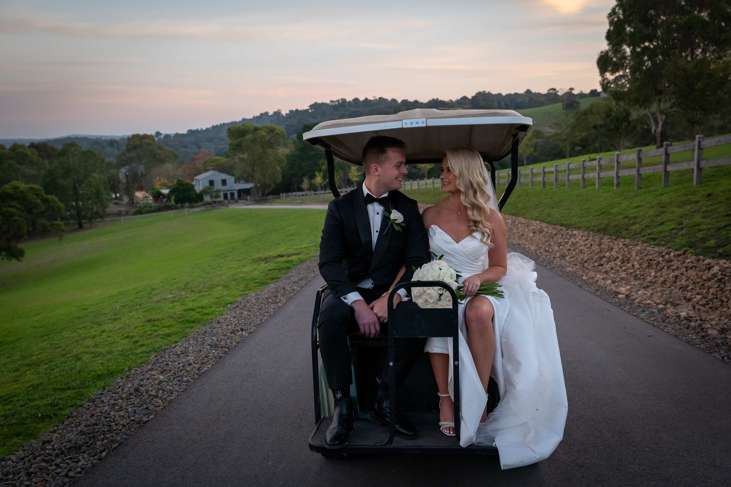 A bride in a white dress and groom in a tuxedo are sitting in a golf cart on a pathway. The bride is holding a bouquet of white flowers. The surrounding landscape is grassy with trees and a house in the distance.