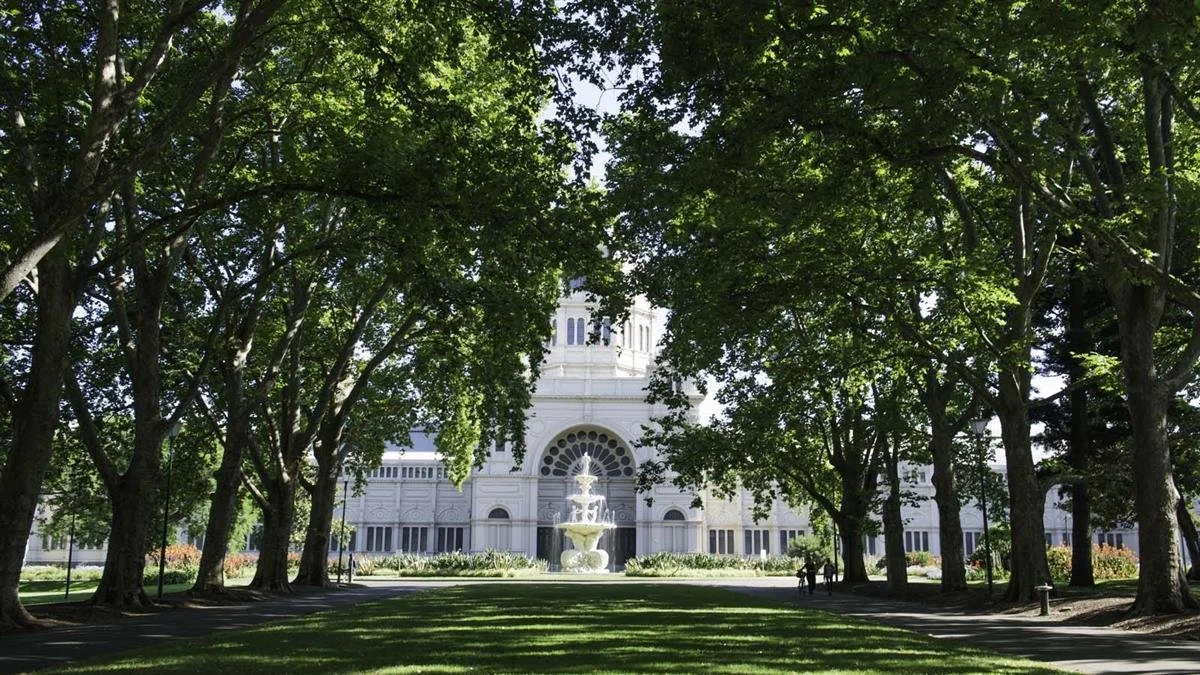 View of a white historic building with a fountain in front, framed by large green-leafed trees in a park.