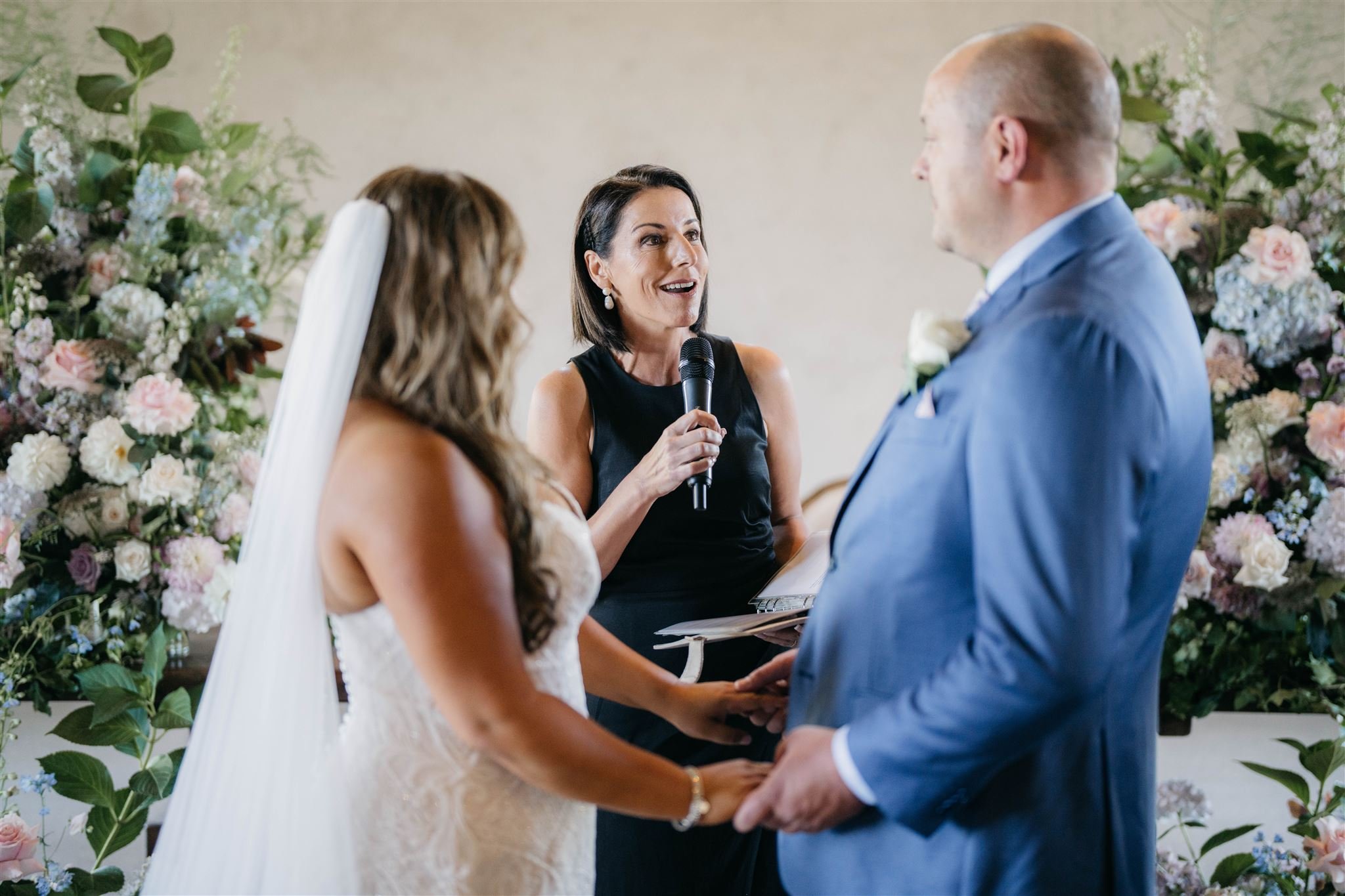 A wedding ceremony with a bride and groom holding hands, while a celebrant speaks into a microphone. They are surrounded by floral arrangements.