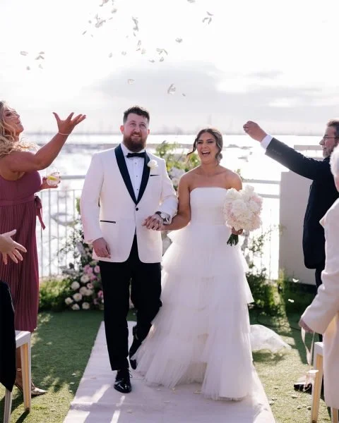 Bride and groom walking down the aisle at their outdoor wedding ceremony, surrounded by friends and family, with confetti in the air and water in the background.