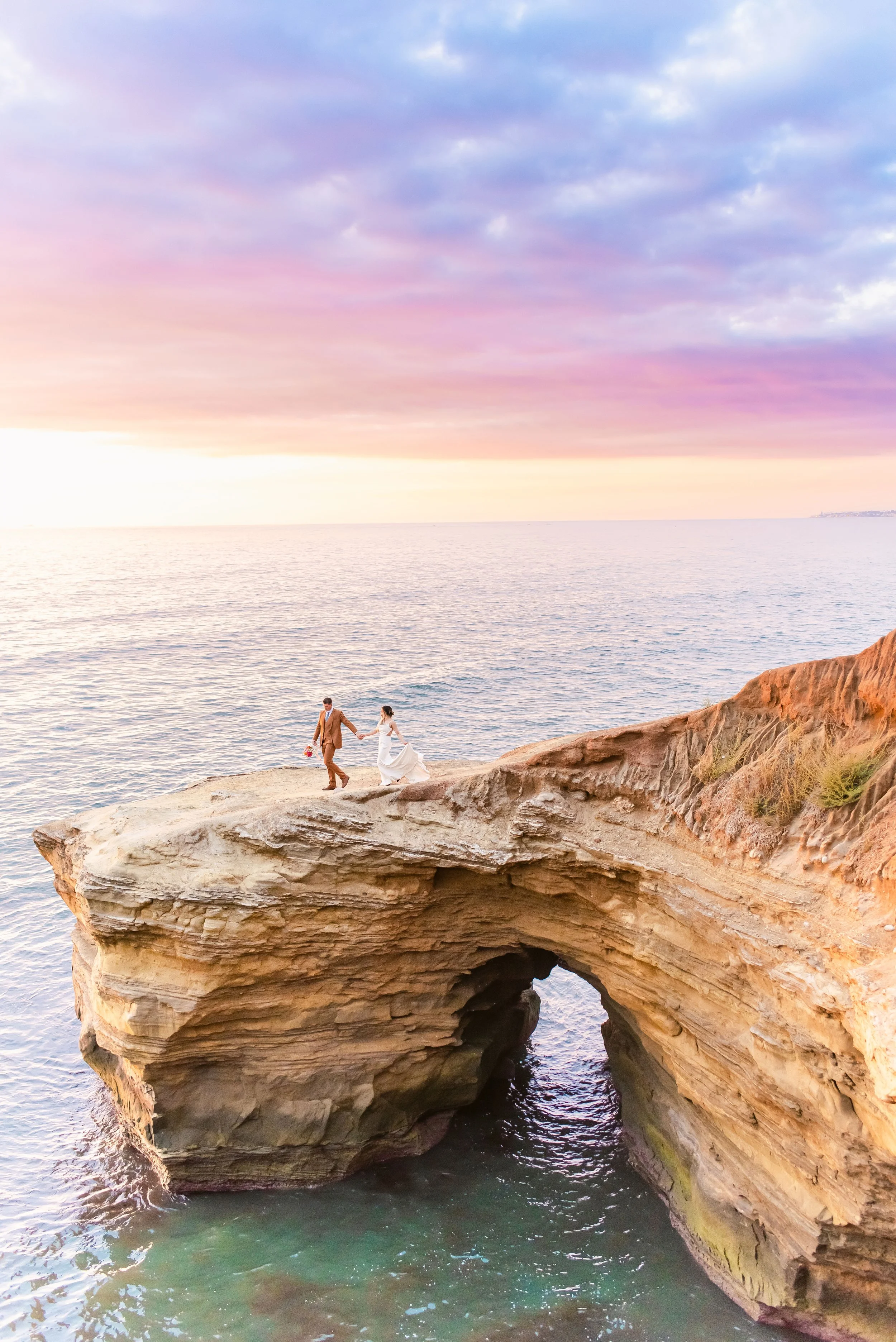 A couple holding hands on a rocky cliff at sunset, with the ocean and colorful sky in the background.