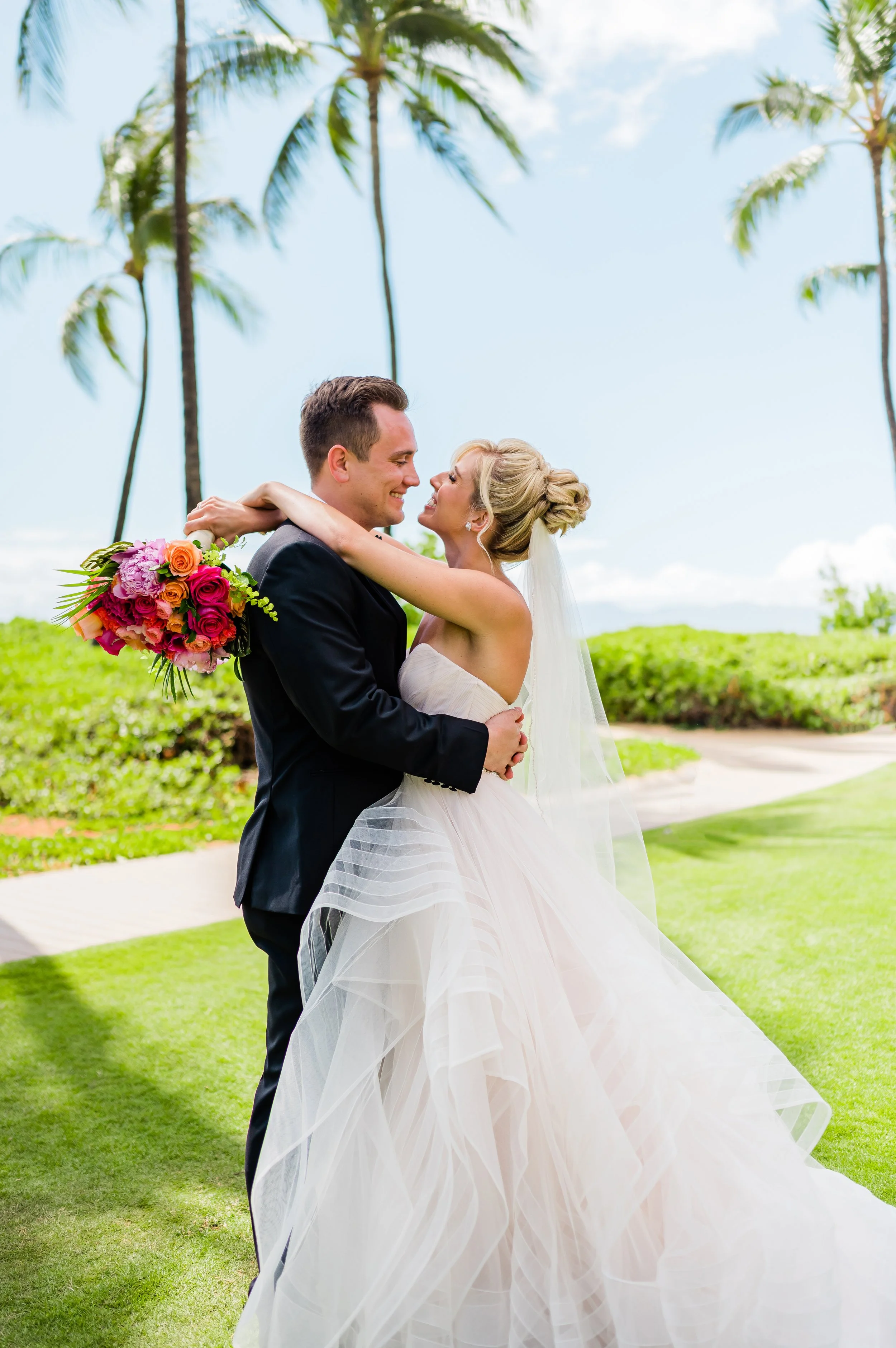 Bride and groom embracing with a bouquet of flowers, palm trees in the background, bright sunny day.