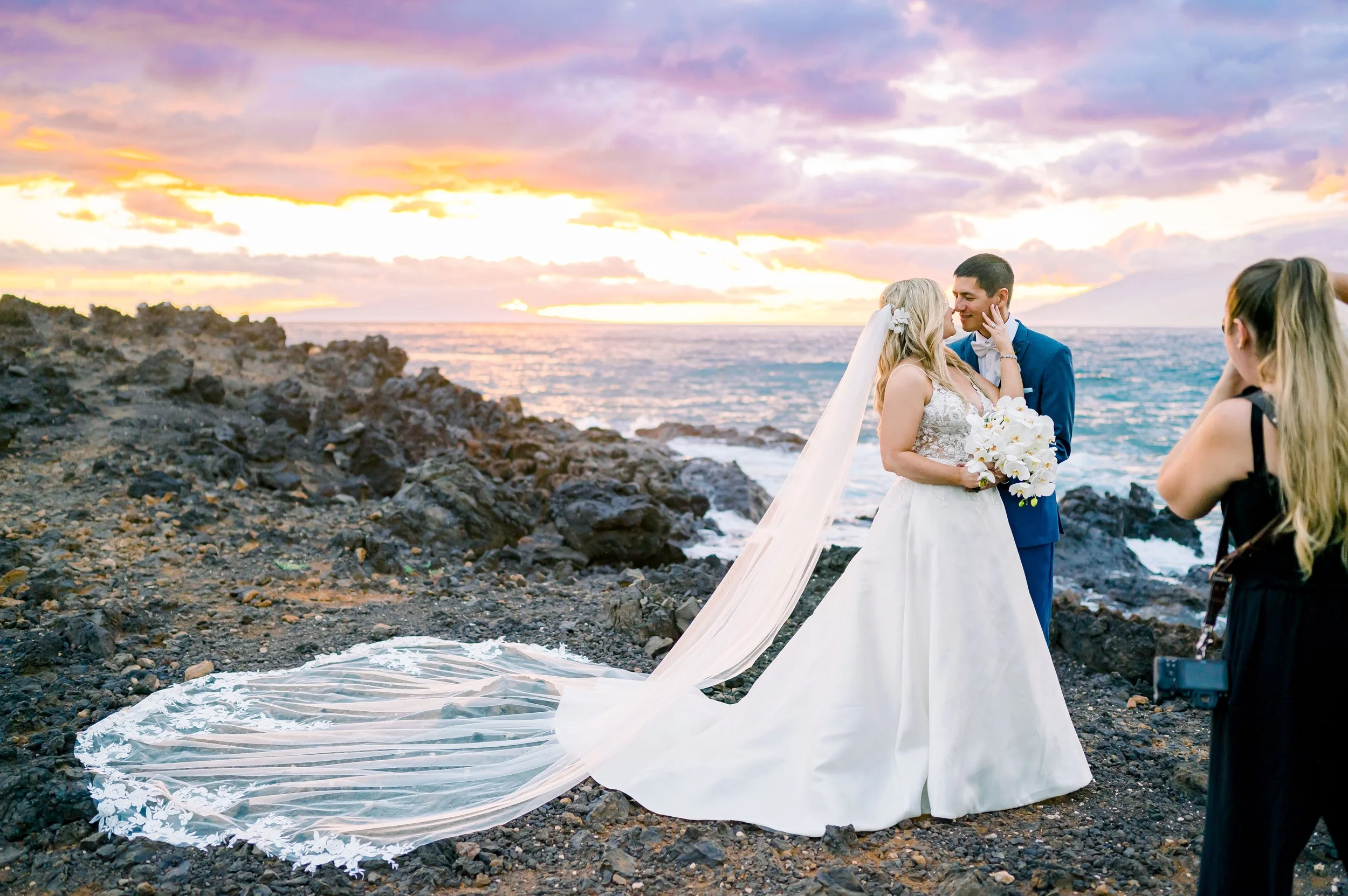 Bride and groom posing near ocean at sunset with photographer capturing the moment. Bride in white gown with long veil, groom in blue suit.