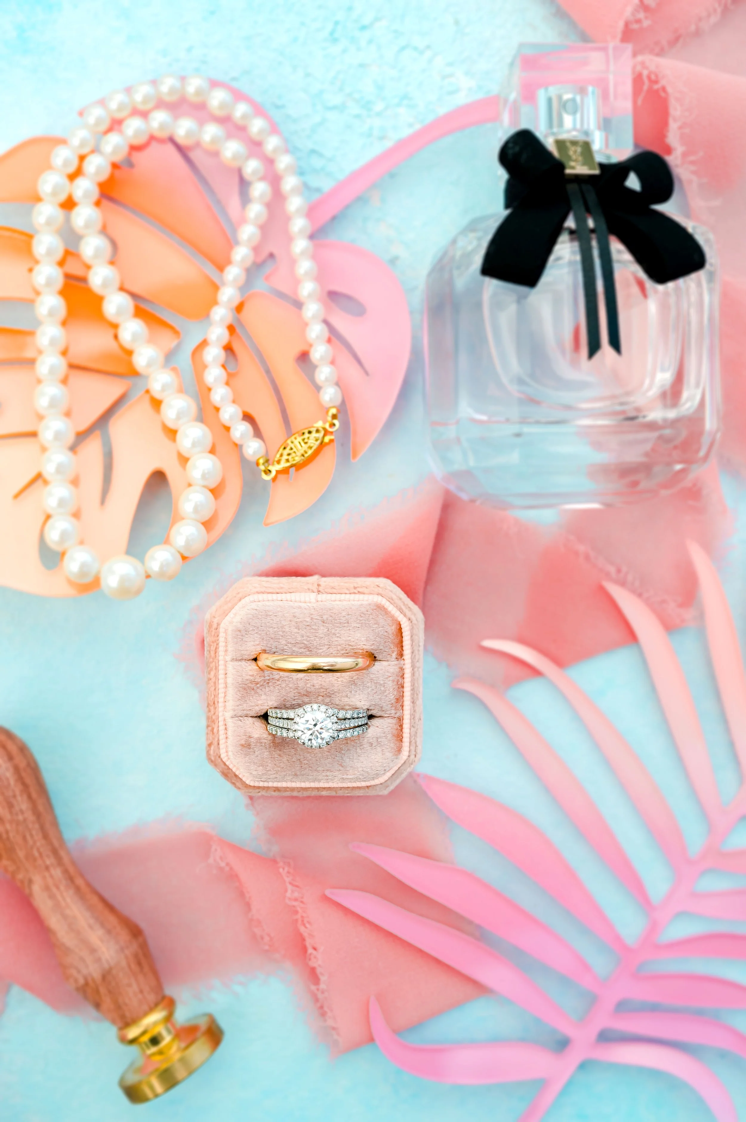 Flat lay of a perfume bottle with a black bow, a string of pearls, a gold wedding band, and a diamond engagement ring in a velvet box, on a pastel blue and pink background with decorative leaves.