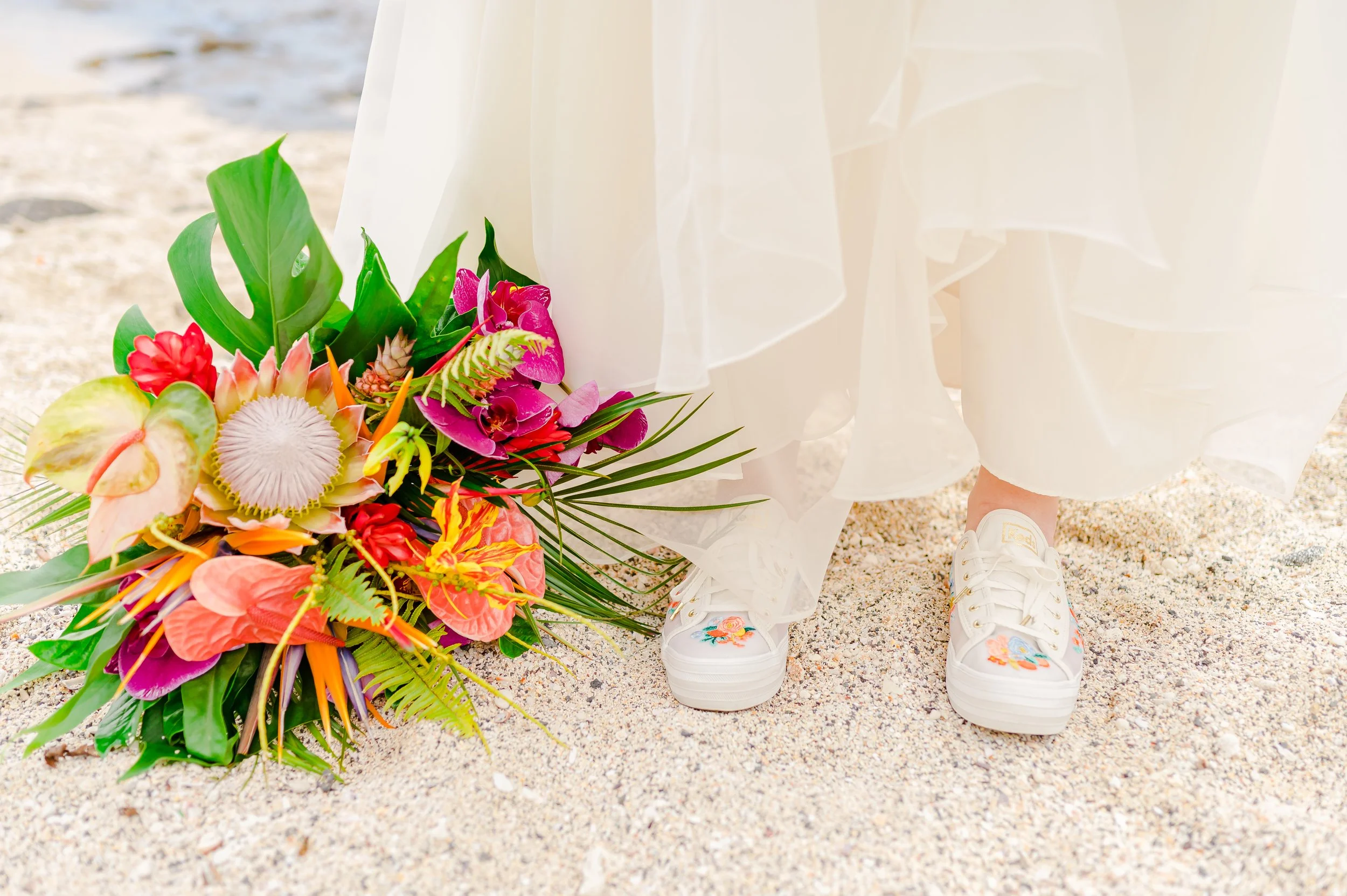 Bride in white sneakers standing on sand next to colorful tropical bouquet.