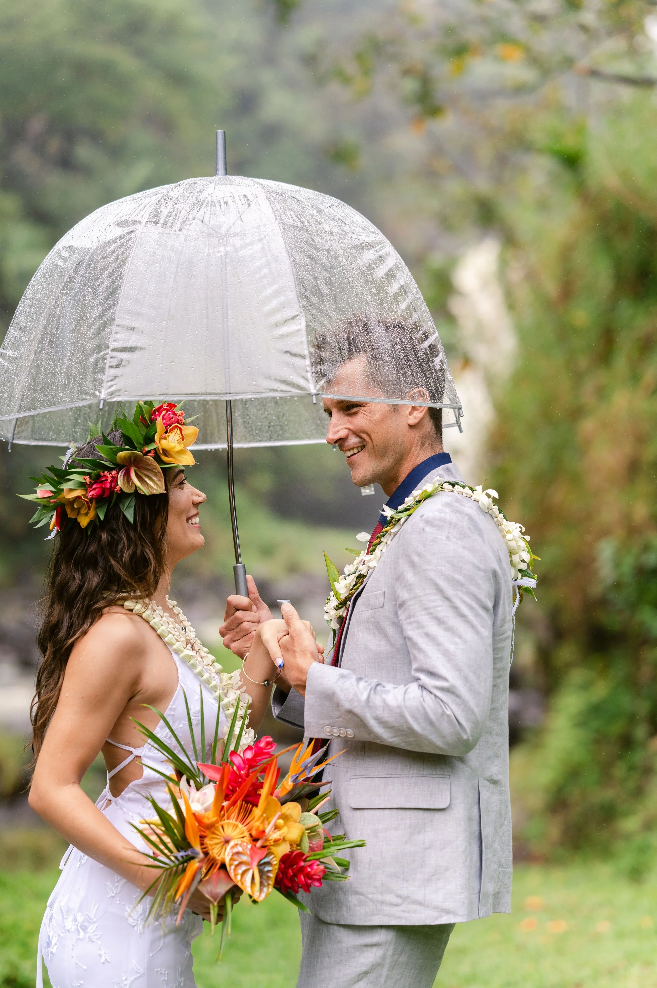 A couple stands under a clear umbrella in an outdoor setting. The woman is wearing a floral crown and a white dress, holding a colorful bouquet. The man is in a light gray suit with a floral lei. They are smiling at each other.