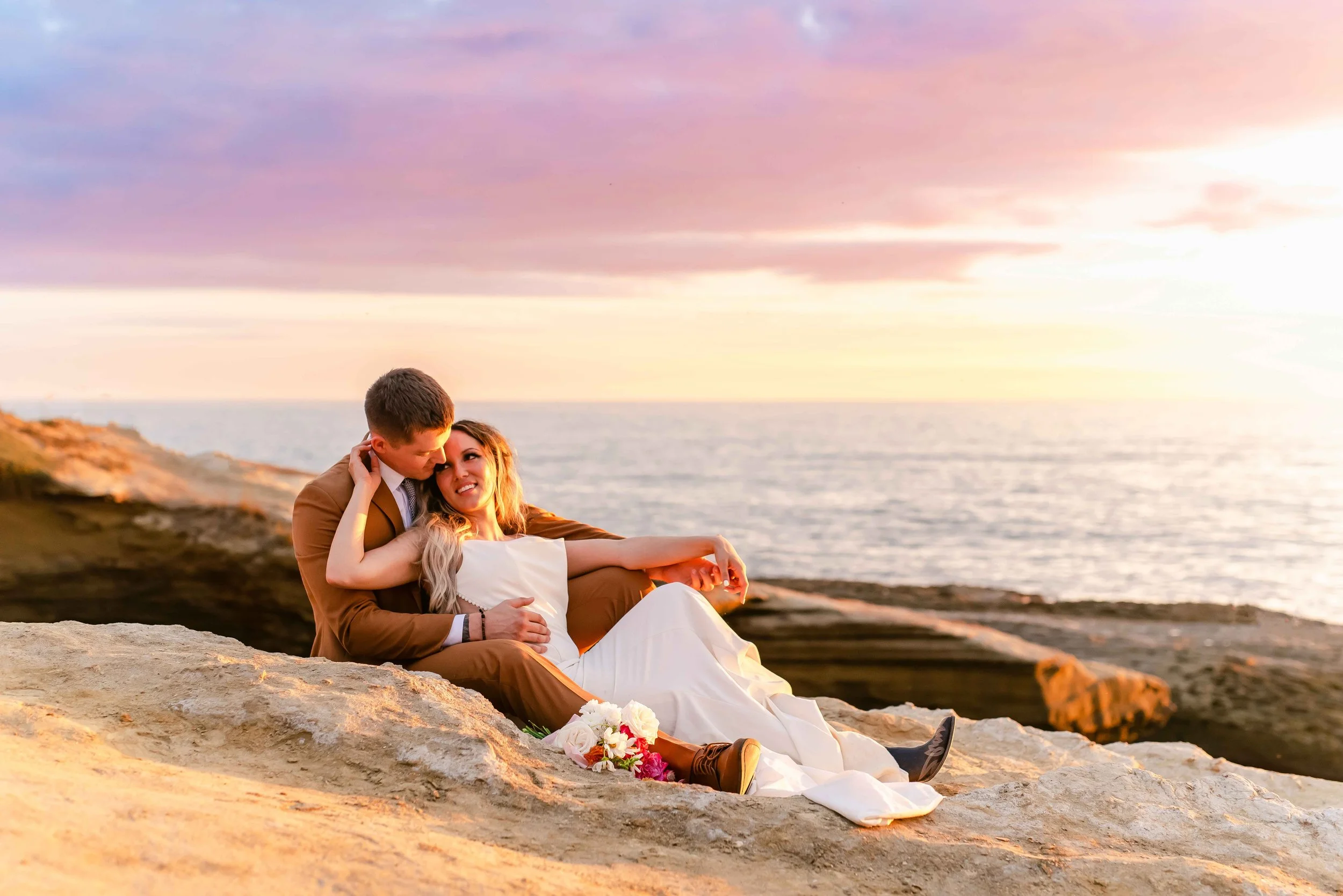 Couple in formal attire sitting on rocky shore during sunset, with ocean in the background.