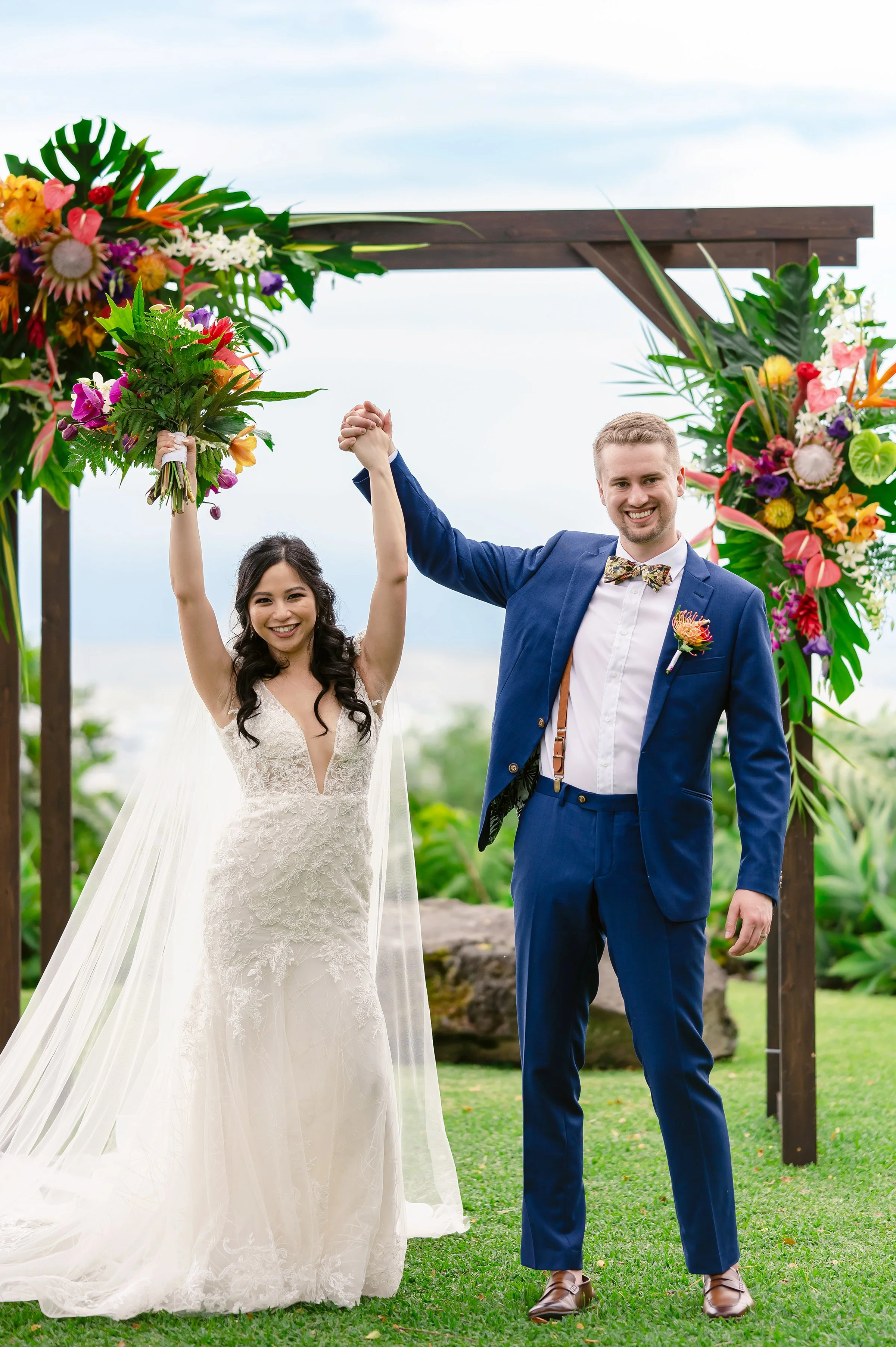 Bride and groom celebrating at an outdoor wedding ceremony, under a flower-decorated arch, with bride holding a bouquet.