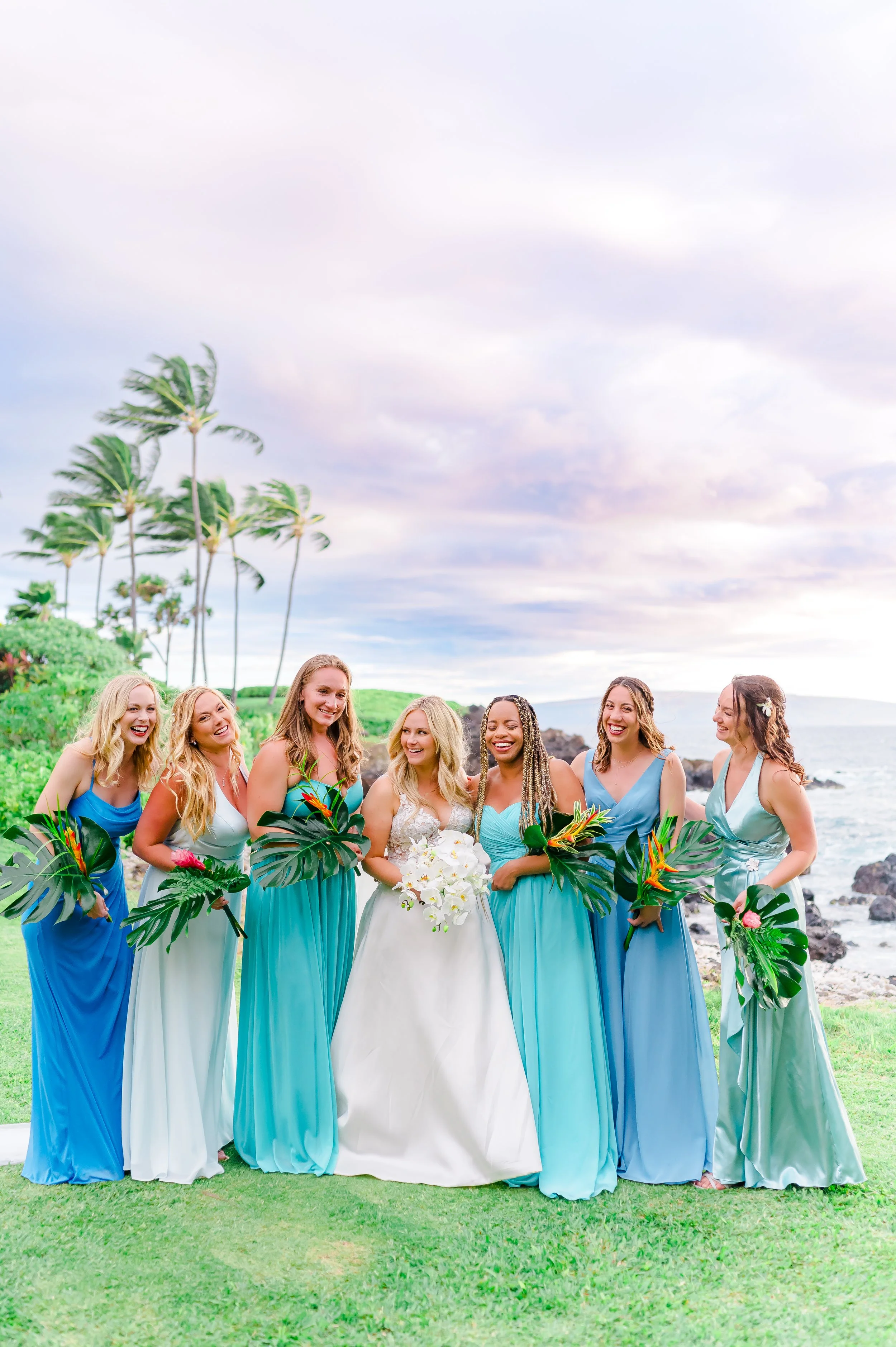 Wedding party with bride and bridesmaids in colorful dresses holding bouquets on a beach setting with palm trees and ocean in the background.