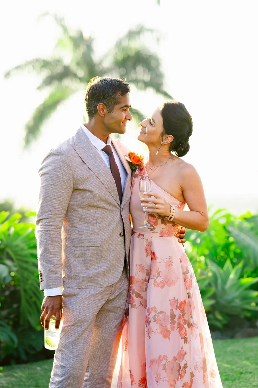 A couple smiling at each other; the man is wearing a light gray suit and tie, the woman is in a floral pink dress, both holding drinks, standing in a garden with palm trees in the background.
