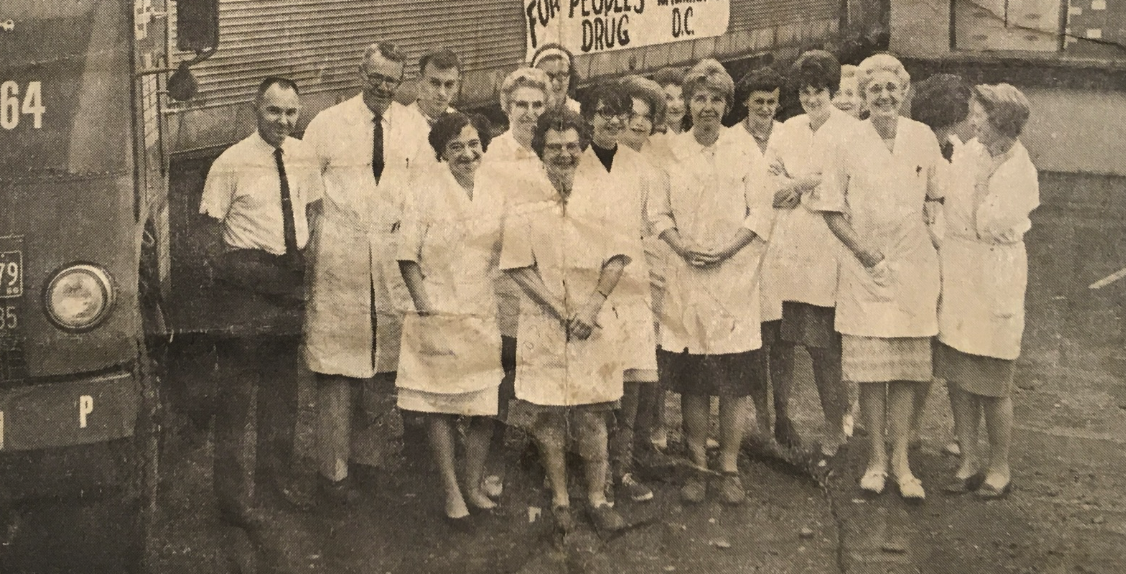 Black and white photo showing a group of women and men standing outside, some wearing white coats or uniforms, in front of a vehicle and a building. There is a sign in the background.