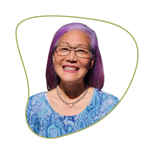 Smiling woman with purple hair, glasses, and a blue patterned blouse, wearing a pearl necklace.