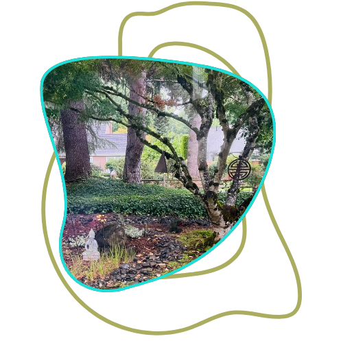 A beautifully landscaped garden with lush greenery, trees, rocks, and a small statue, viewed through a uniquely shaped window or frame.