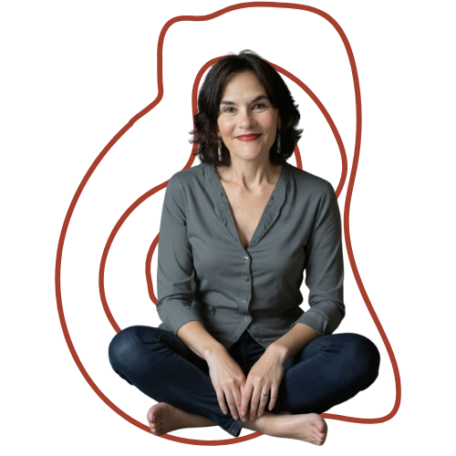 A woman with shoulder-length dark hair, wearing a gray button-up shirt and blue jeans, sitting cross-legged on the floor and smiling.