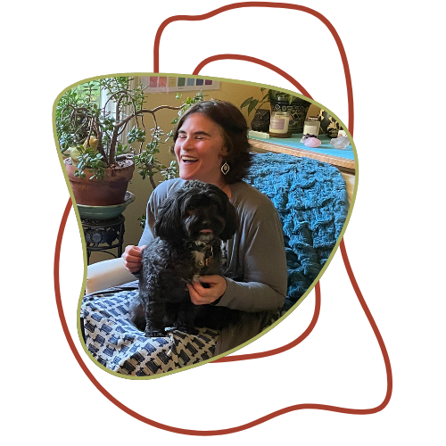 A woman sitting on a couch holding a black dog, smiling in a cozy living room with plants and a blue blanket.