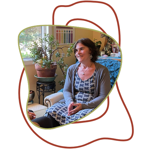 A woman sitting on a cream-colored armchair with a floral dress and gray cardigan, smiling inside a cozy room with plants and colorful decor.