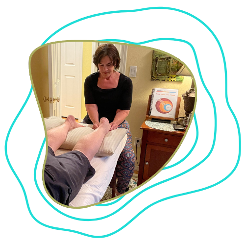 A woman performing a foot massage on a person lying on a massage table in a room with a dresser, a lamp, and a computer monitor displaying a colorful chart.
