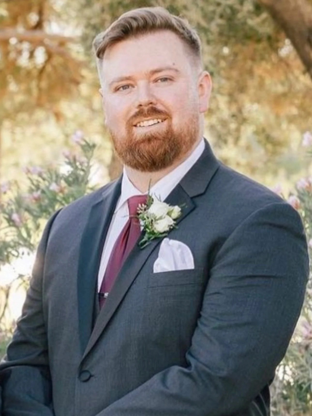 A man with a red beard and short, styled hair, wearing a black suit, white shirt, red tie, and a white boutonniere, smiling outdoors with trees and flowers in the background.