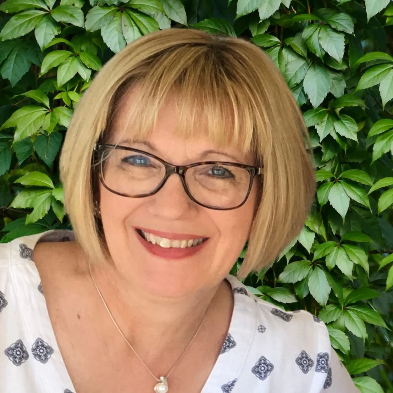 Woman smiling outdoors with blonde bob, wearing a white short sleeve blouse, green leaves in background.