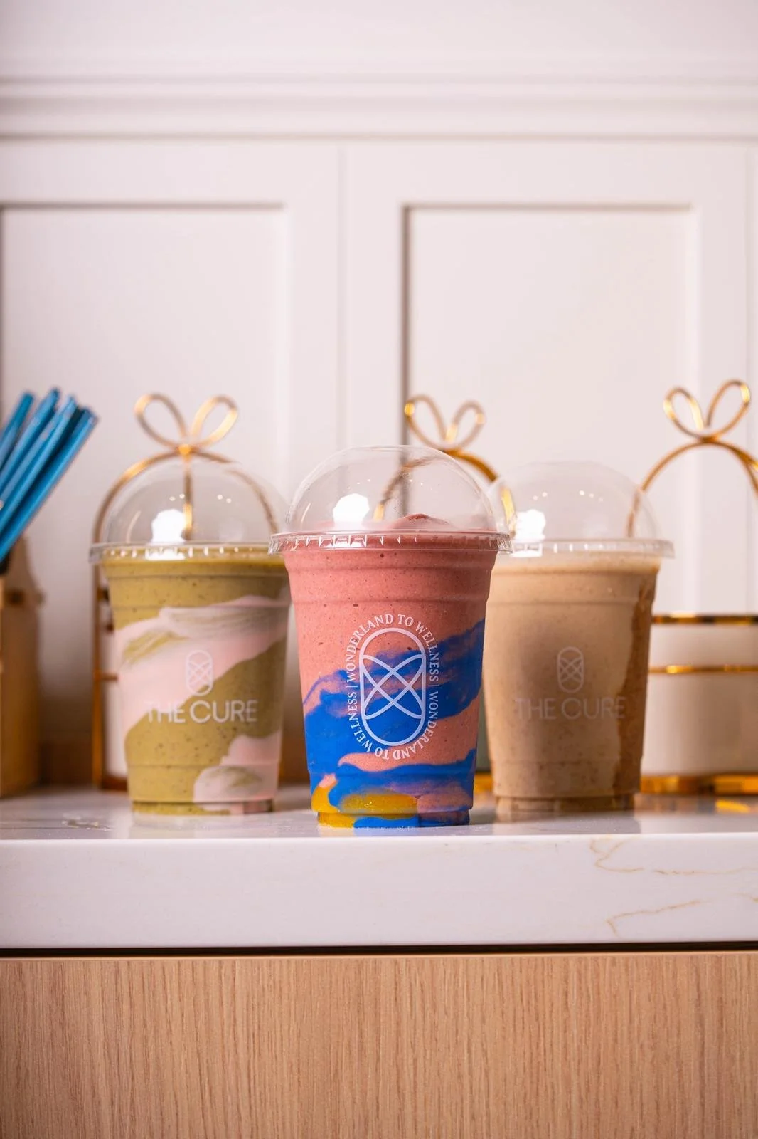 Three colorful smoothies in clear plastic cups with domed lids on a white counter, with a white wall and gold wire racks in the background.