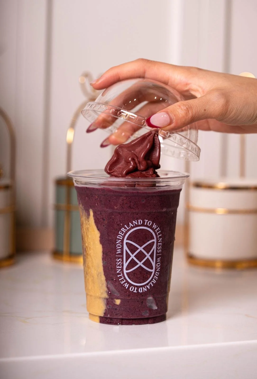 A hand lifting the lid off a dark purple smoothie in a clear plastic cup with gold accents in the background. The smoothie appears to be chocolate or berry flavored, with some yellow and purple marbling. The cup has a circular logo with the words 'WONDERLAND TO WELLNESS' printed around it.