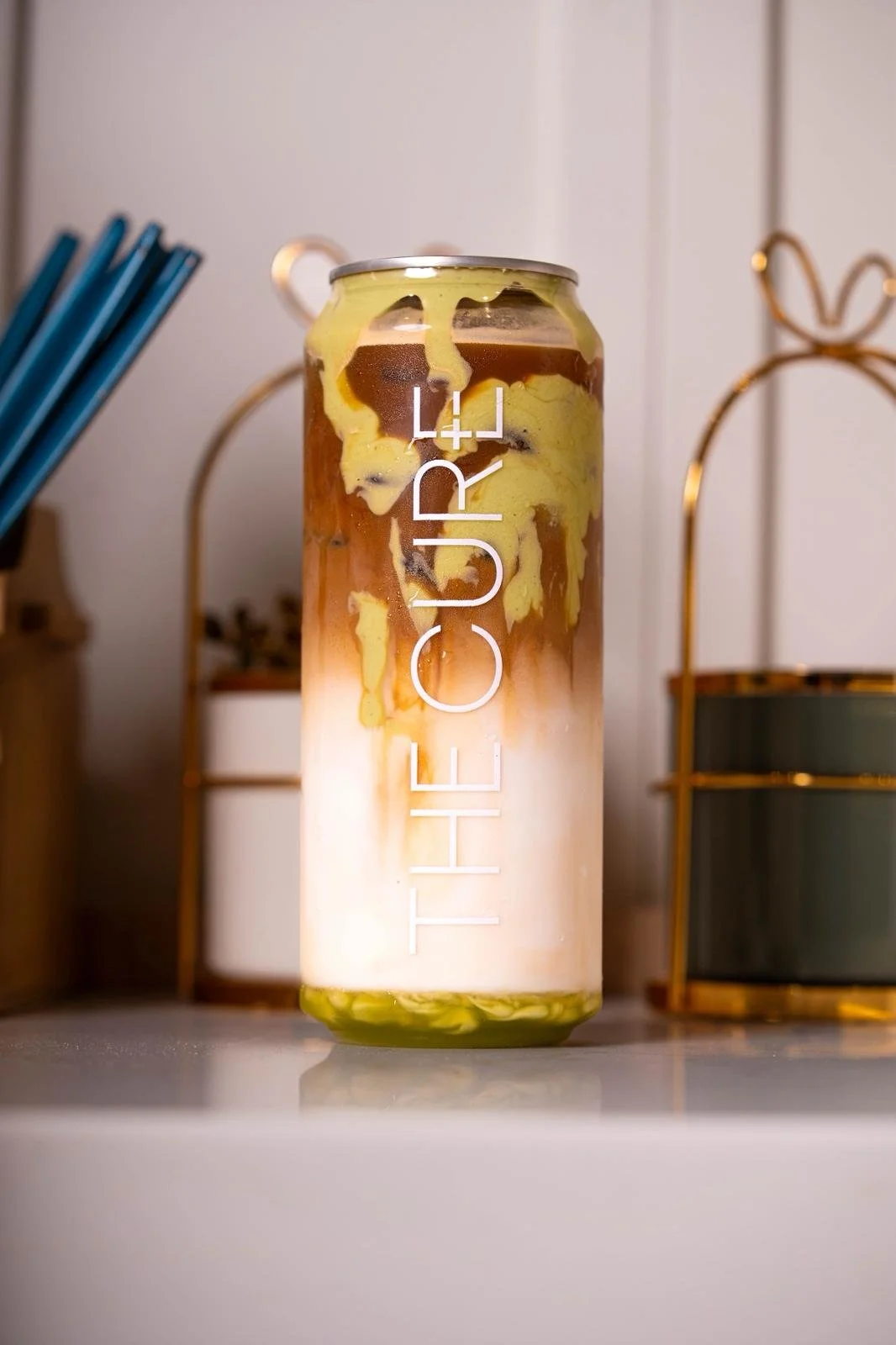 A layered iced coffee drink in a tall glass with the word "THE CURE" written on it, showcasing swirls of milk, coffee, and yellow syrup or flavoring.